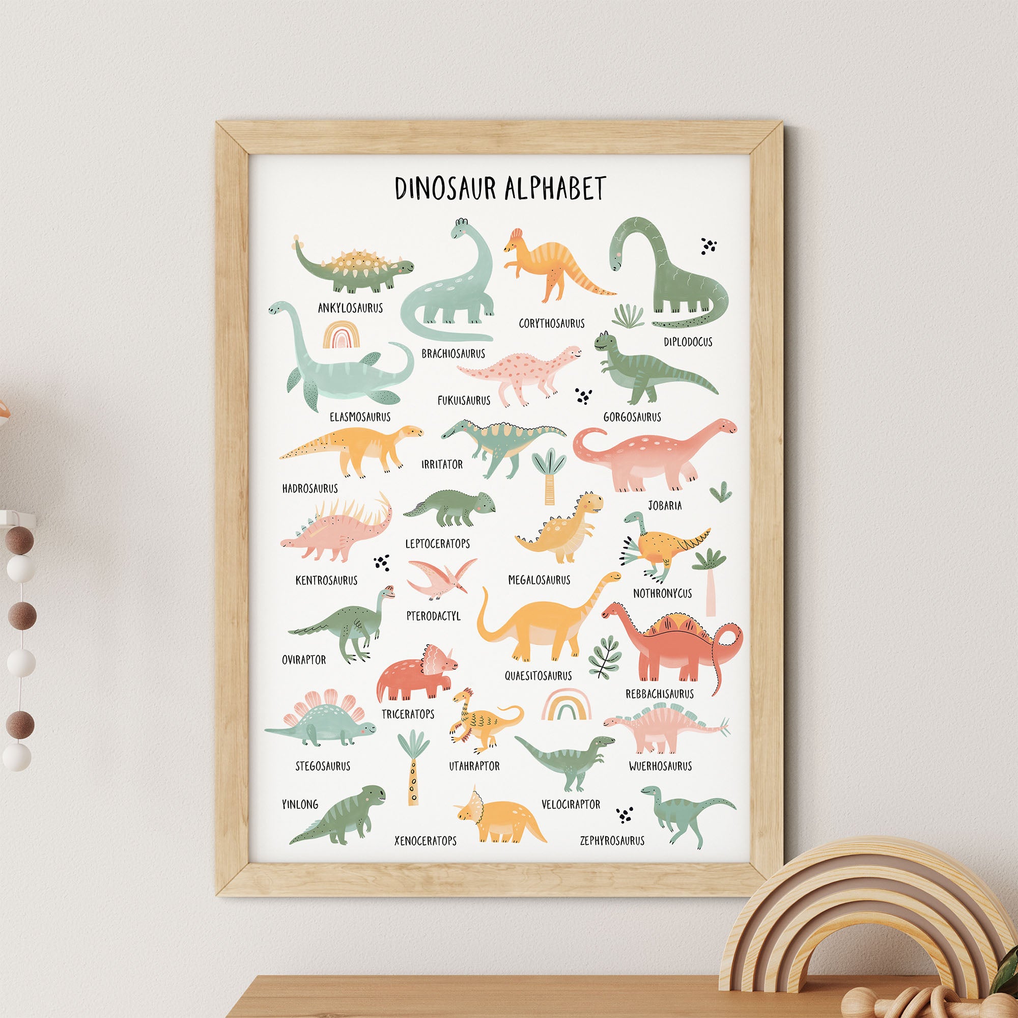 Framed poster titled Dinosaur Alphabet features illustrated dinosaurs, each labeled with names from A to Z, displayed in rows on a white background and hung on a light-colored wall.