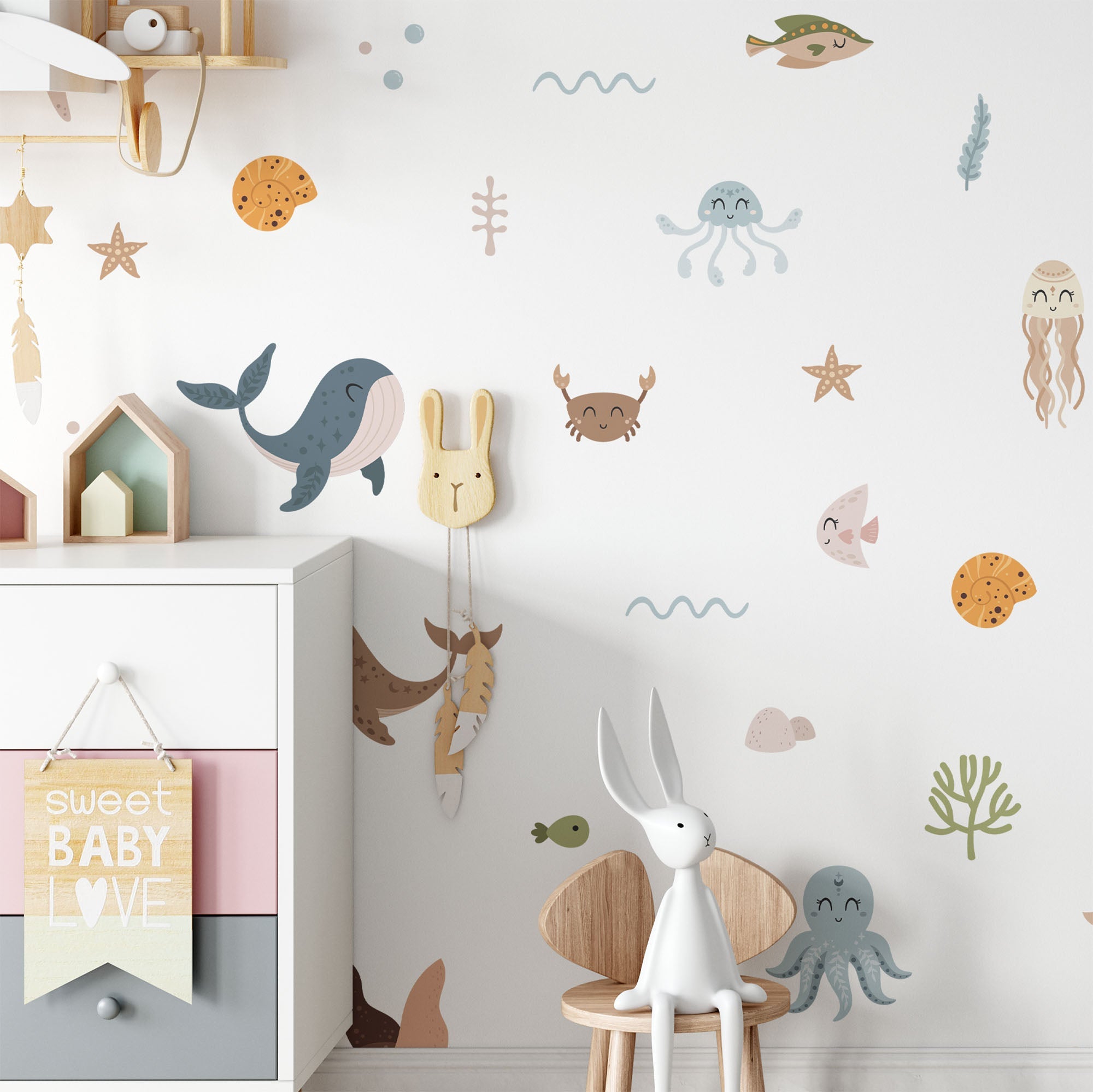 A nursery room with sea animal wall decals, a white dresser with pastel decor, a bunny chair, and a sign that says “sweet BABY LOVE.” There are plush toys and hanging decorations.
