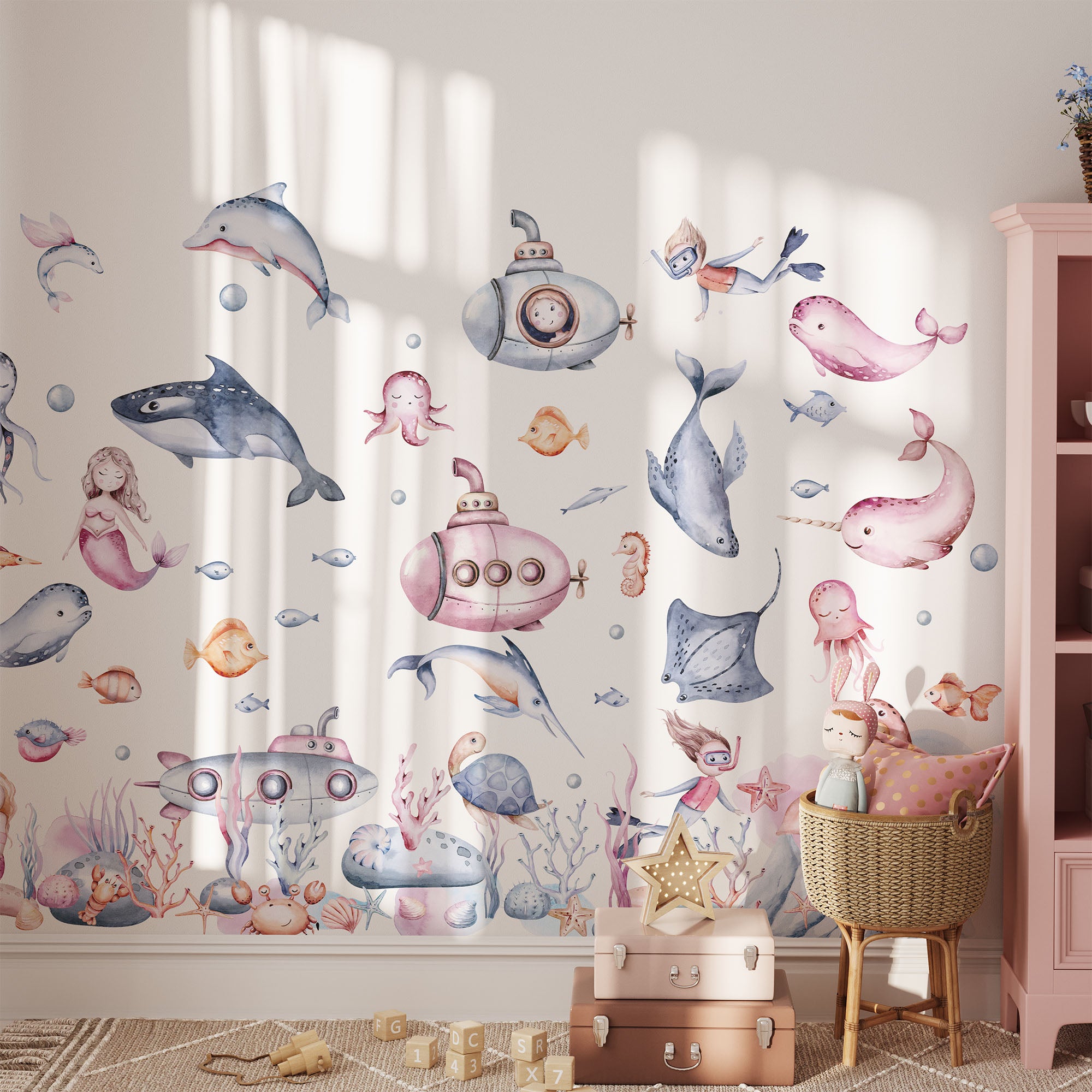 Children’s room with pastel undersea-themed wall decals showing whales, dolphins, submarines, fish, a mermaid, and coral. Plush toys and a pink shelf decorate the cozy space.