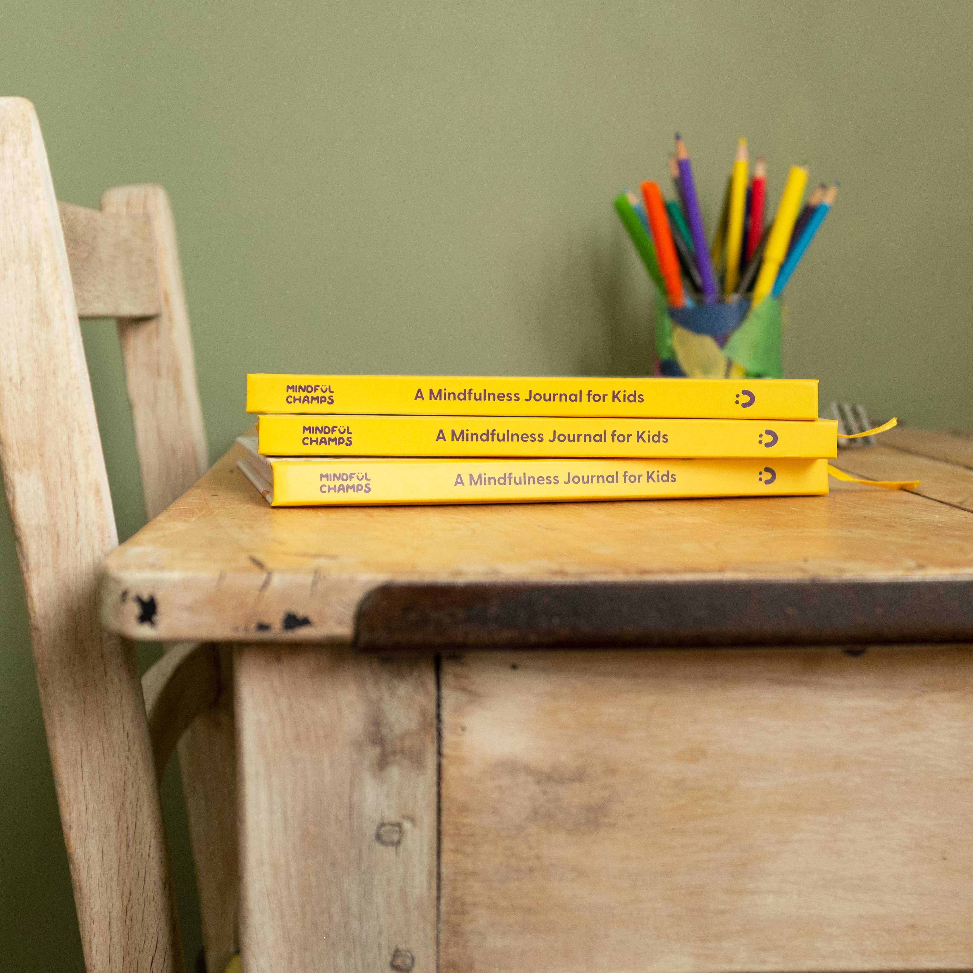 Three yellow “My Mindful Me Journal” books by Mindful Champs are stacked on a wooden desk to support children’s mental health. A wooden chair is beside the desk, and a green cup with colorful pencils sits at the back. The wall is light green.
