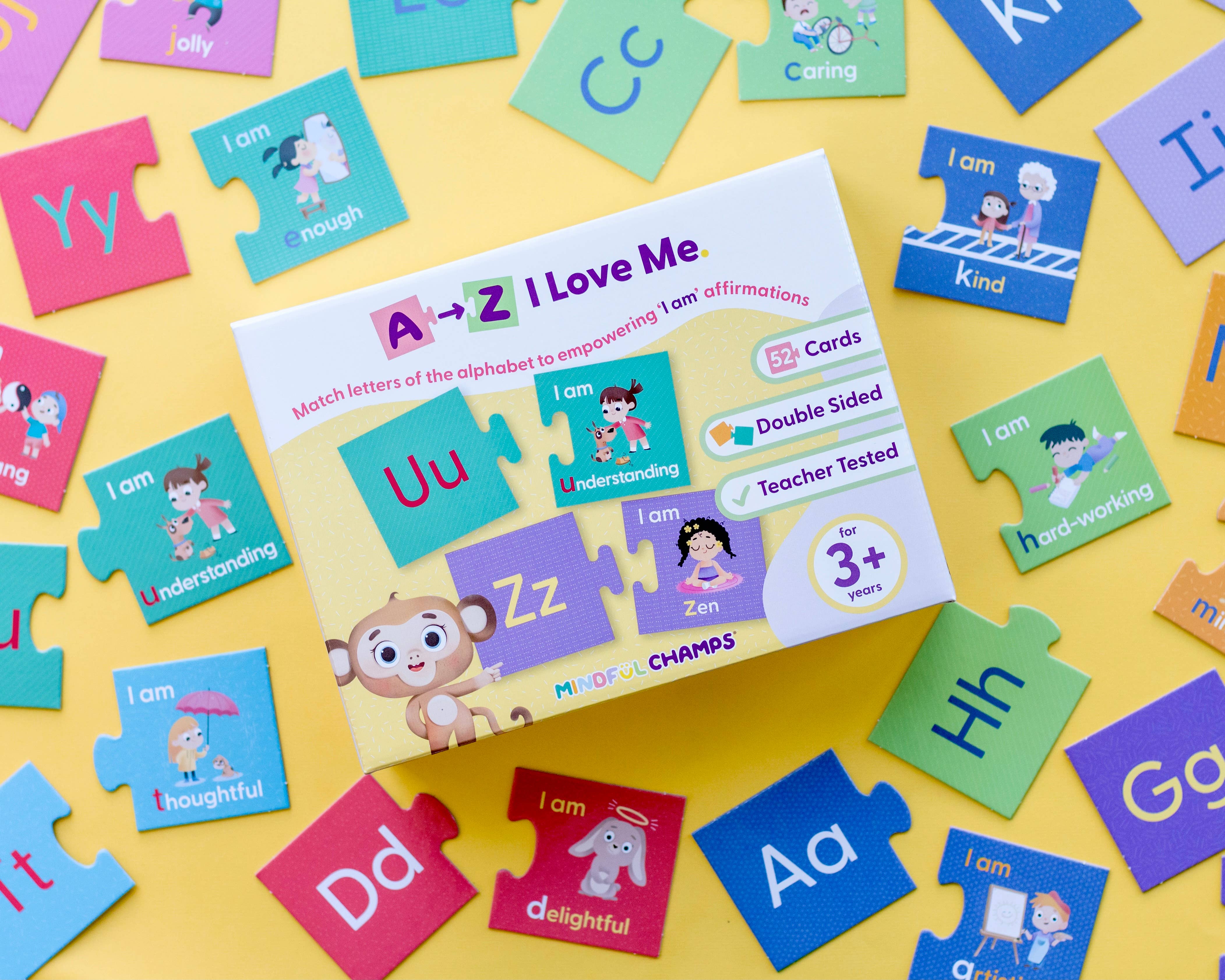 The Mindful Champs 'A-Z I Love Me' Affirmation Puzzle sits on a yellow surface, surrounded by letter pieces with positive messages like “I am understanding.” The box displays a cartoon monkey and encourages kids’ self-esteem through alphabet matching.