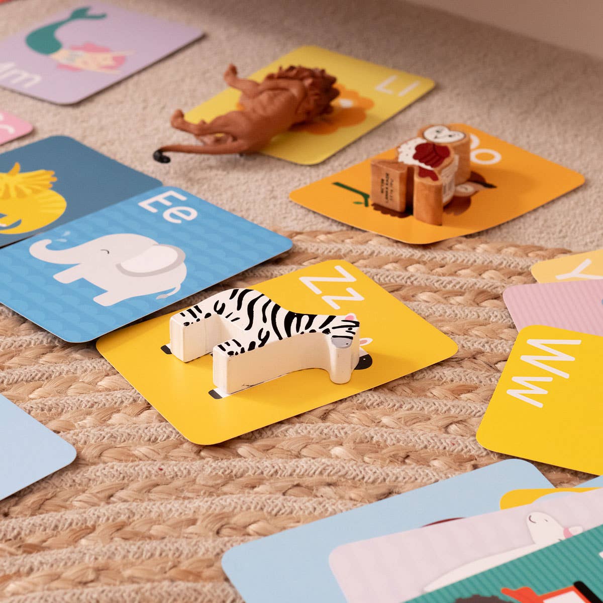 Priya & Peanut's Alphabet Flash Cards | Learn The Alphabet Easily are laid out on a woven rug, each featuring a toy figure—like a zebra for Z, lion for L, elephant for E, and owl for O—for fun educational play.