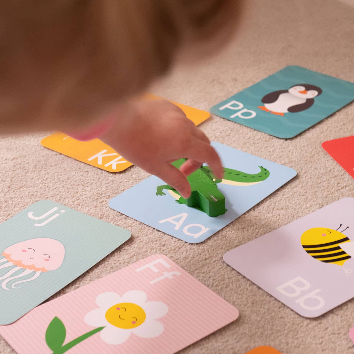 A child’s hand places a green toy alligator on a blue “Aa” Priya & Peanut Alphabet Flash Card from the Alphabet Flash Cards | Learn The Alphabet Easily set, surrounded by other colorful animal-themed cards on a carpet.