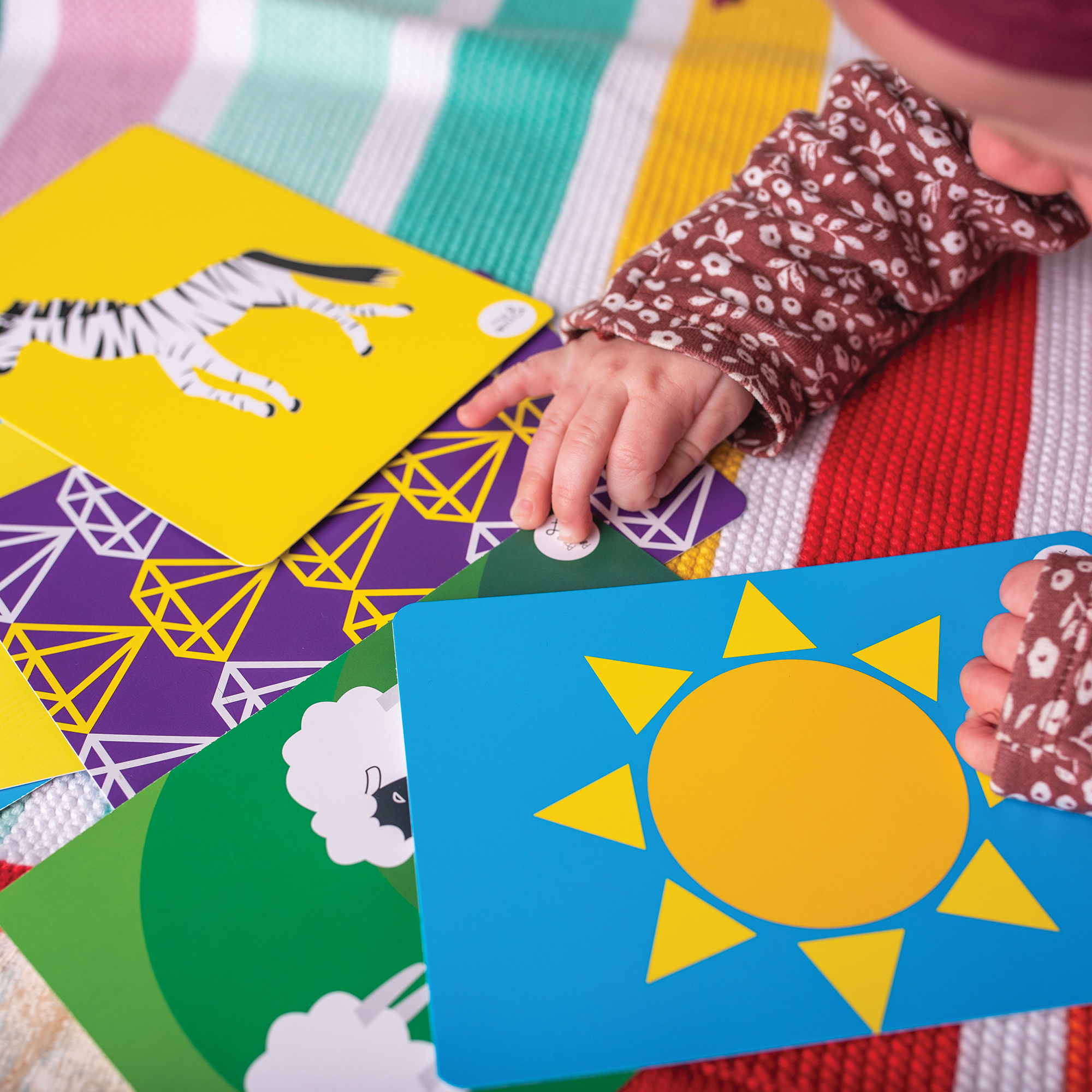 A baby reaches for Priya & Peanut’s Colour Sensory Flash Cards For Babies (3+ Months), a sensory toy with simple, high-contrast illustrations—sun, zebra, and sheep—touching the card with the sun design on a striped blanket.