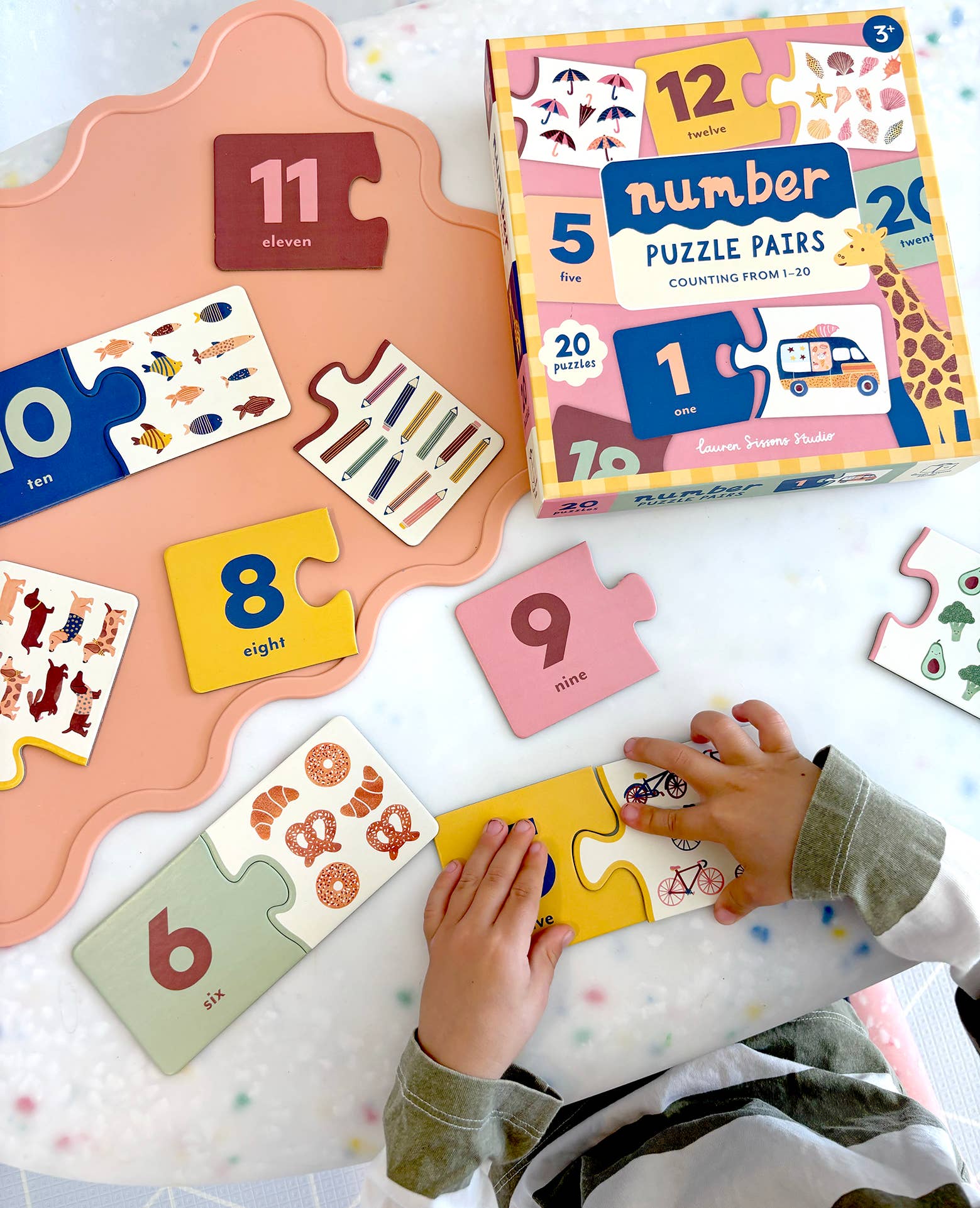 A child connects colorful pieces from Lauren Sissons Studio’s "Number Puzzle Pairs - Kids Toys Learning Game Educational," building counting skills. Mismatched pieces and a box with numbers and animal illustrations are also on the table.