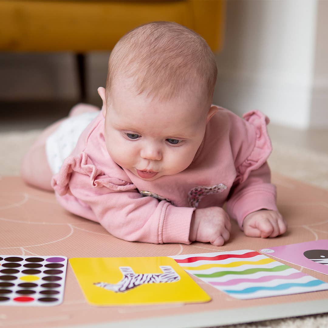 A baby in a pink outfit lies on their stomach on a play mat, engaging with Priya & Peanut's Colour Sensory Flash Cards For Babies (3+ Months), a sensory toy designed to support early development.