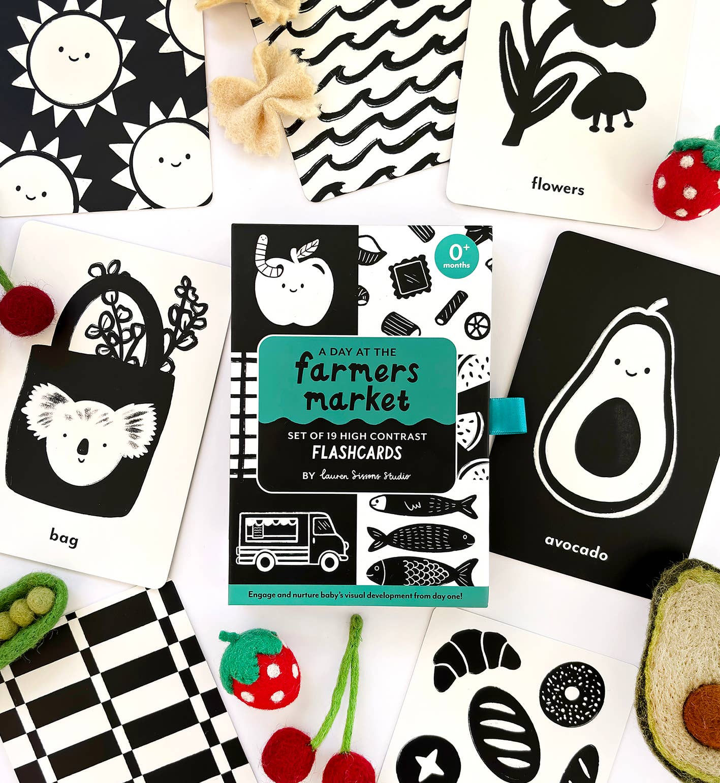 Lauren Sissons Studio's High Contrast Flashcards for Baby – Sensory Newborn Gift are arranged with felt strawberry toys, featuring simple black-and-white images like avocado, flowers, a bag, and fish to aid newborn visual development.