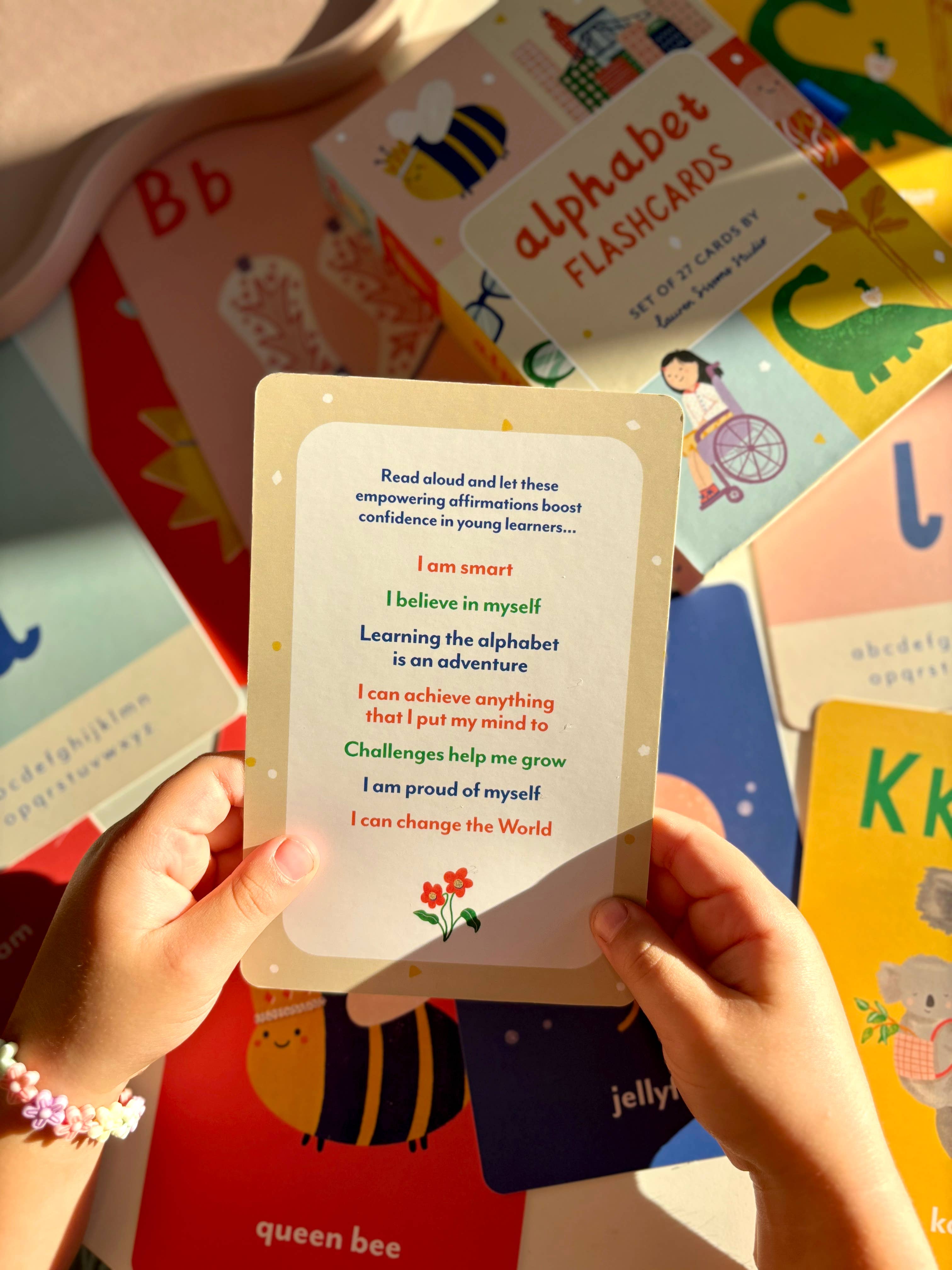 A child holds a Lauren Sissons Studio A-Z Illustrated Alphabet Flashcard with motivational phrases, surrounded by colorful learning cards—a sunlit, joyful scene highlights these educational kids’ toys.