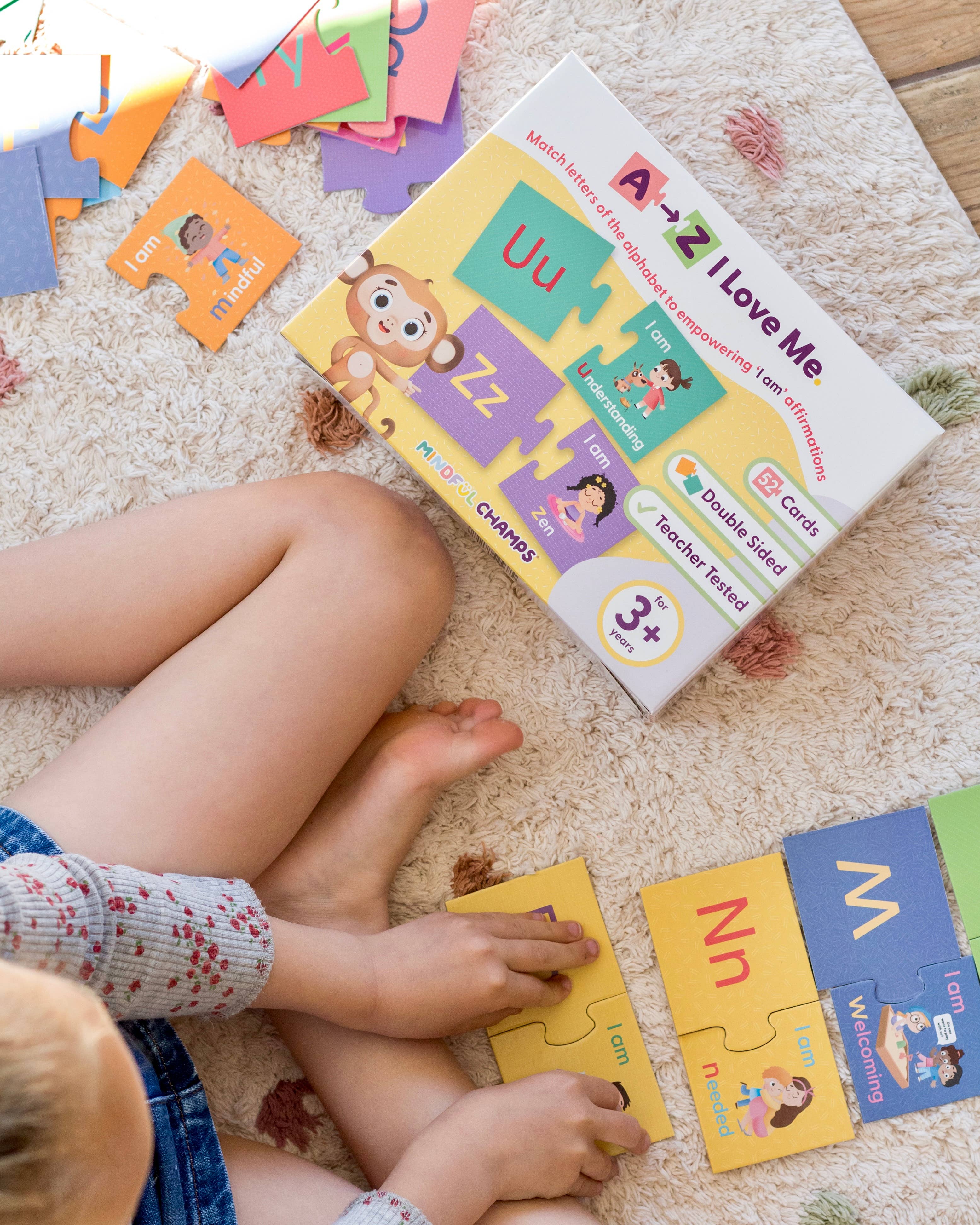 A child sits on a carpet playing with colorful pieces from the Mindful Champs 'A-Z I Love Me' Affirmation Puzzle, an alphabet matching game with affirmation cards that encourages a positive self-image. The game box is also visible.