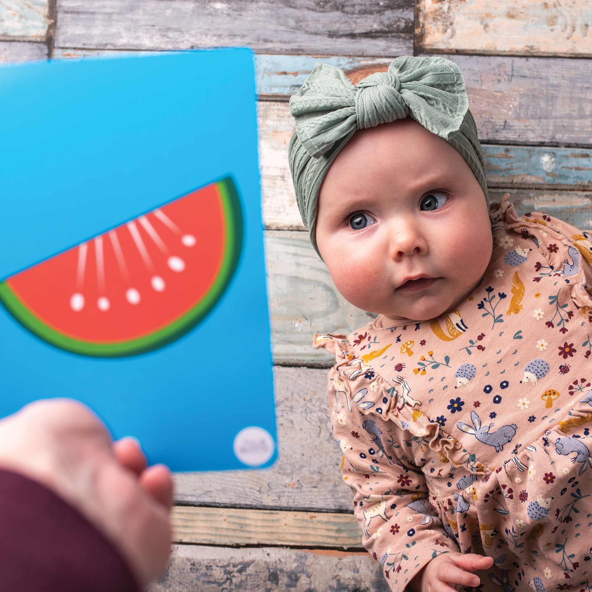 A baby in a green headband and floral outfit lies on a wooden floor, gazing at Priya & Peanut Colour Sensory Flash Cards For Babies (3+ Months)—a sensory toy designed to encourage baby development with contrasting colours.