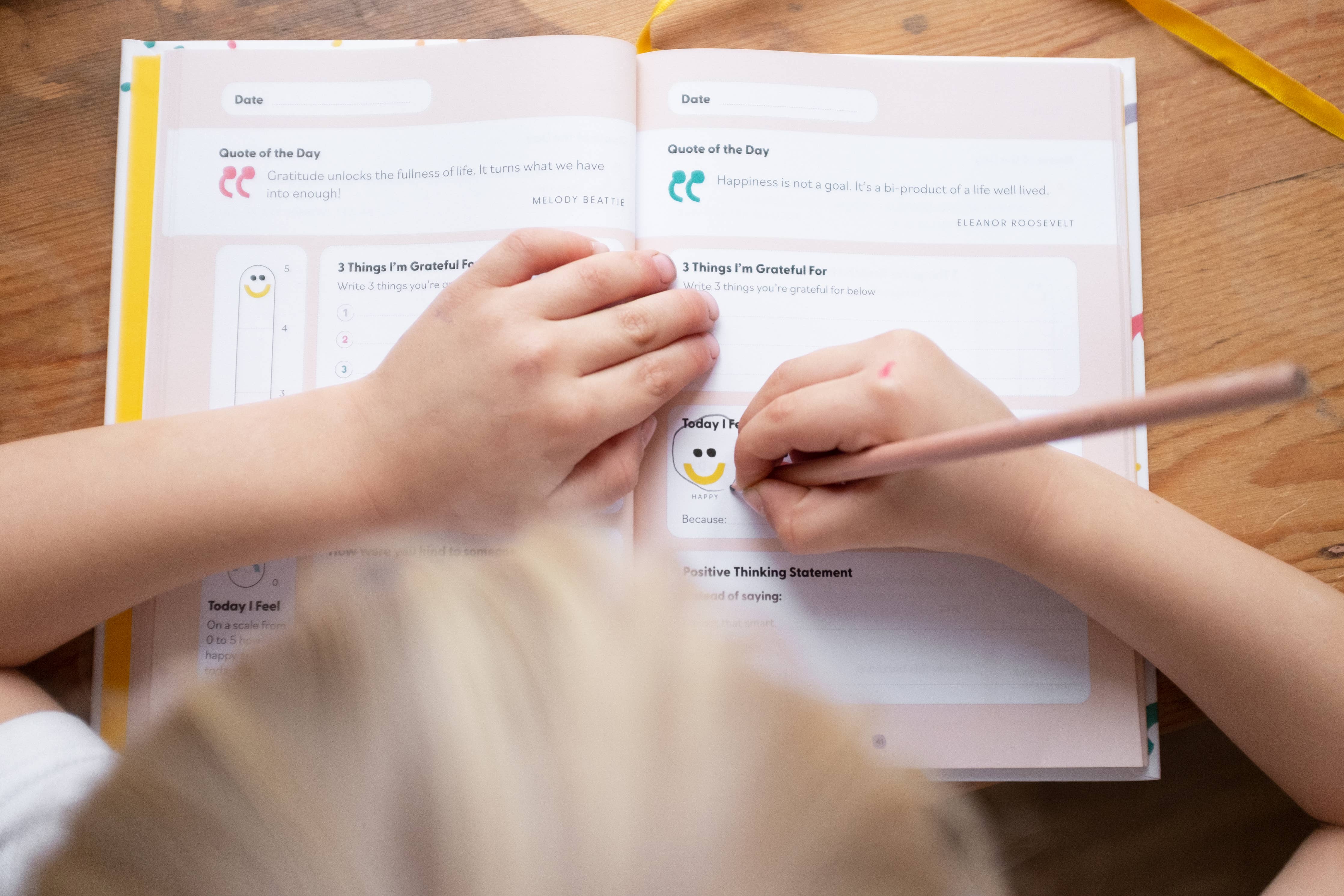 A child writes with a pencil in the My Mindful Me Journal by Mindful Champs, filling out gratitude prompts at a wooden table as another child's hand rests gently on the open page.
