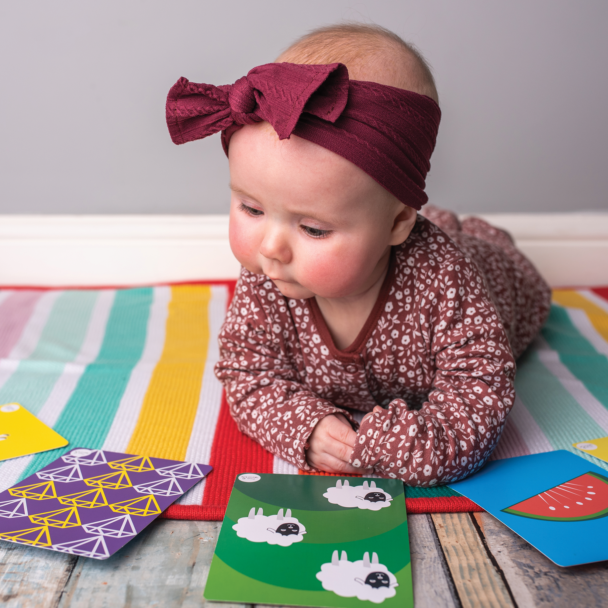 A baby in a maroon headband and patterned outfit lies on a colorful mat, exploring Priya & Peanut's Colour Sensory Flash Cards For Babies (3+ Months) to boost early development with bright images like sheep, shapes, and watermelon.