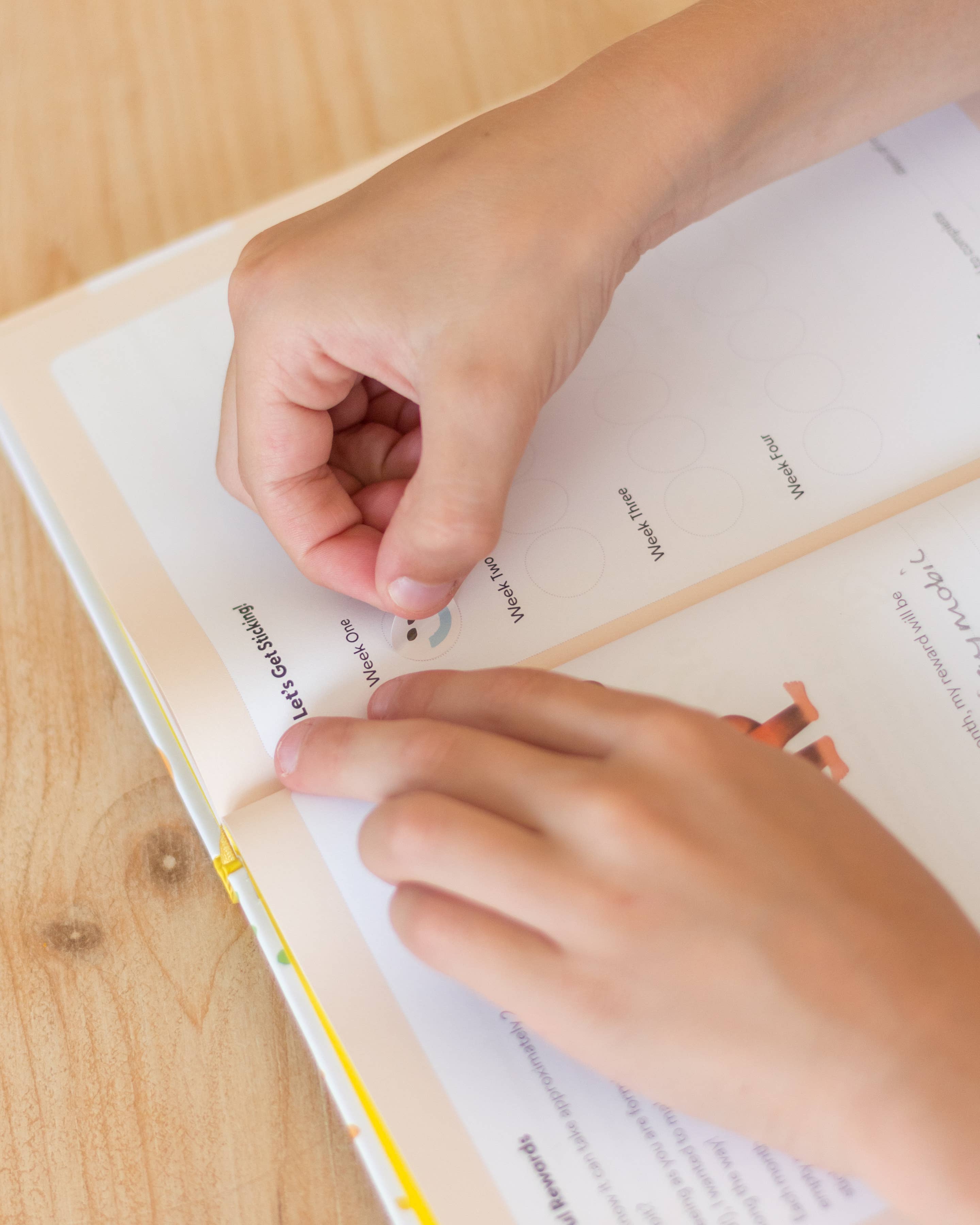 A person’s hands place a round sticker onto a page in an open My Mindful Me Journal by Mindful Champs, which lies on a wooden surface. The page includes printed text and circles for stickers.