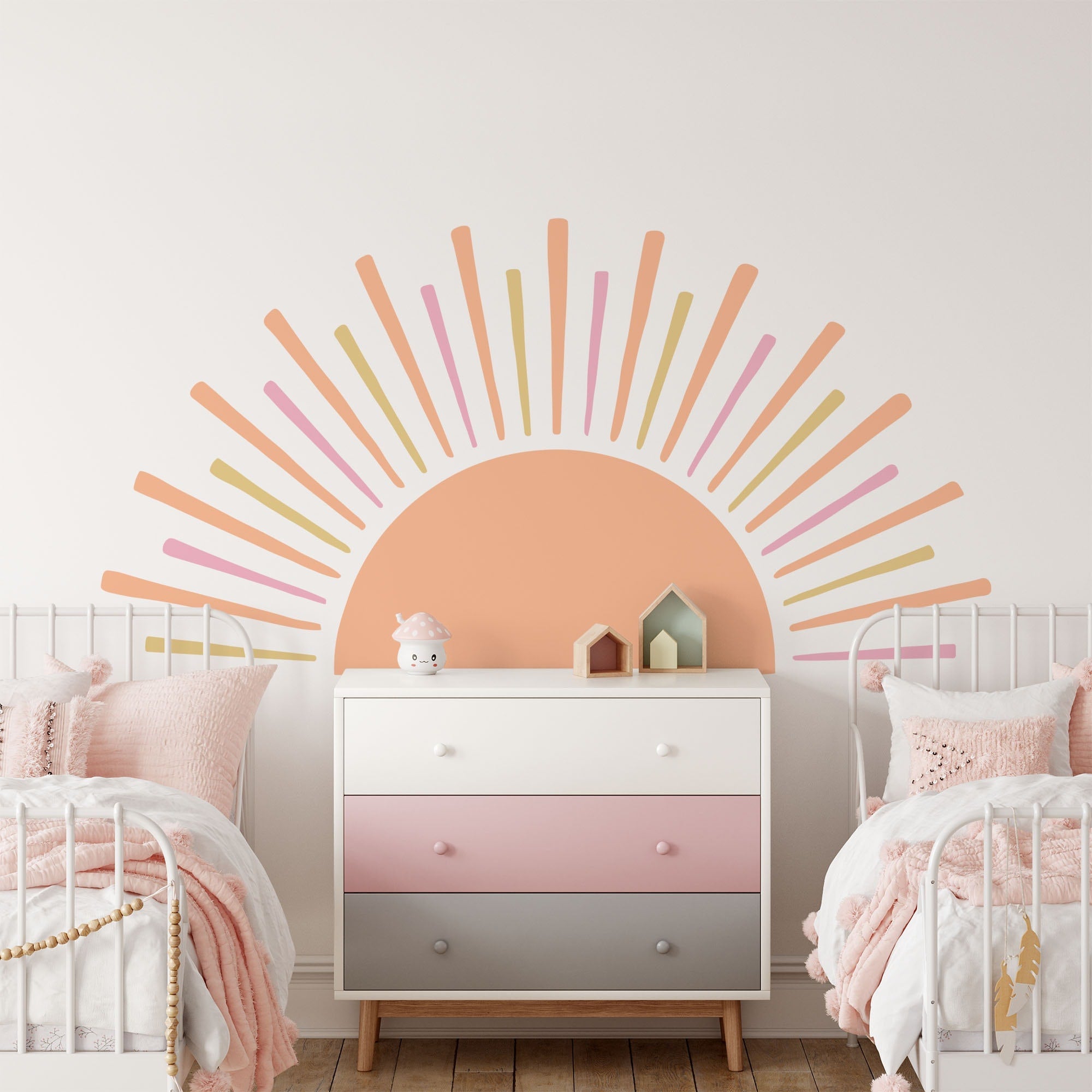 A kids room with two white beds featuring pink bedding, a pastel dresser in the center adorned with My Stores Boho Sun Sticker depicting an orange, pink, and yellow sun. Plush toys and small houses complete the charming nursery decor.