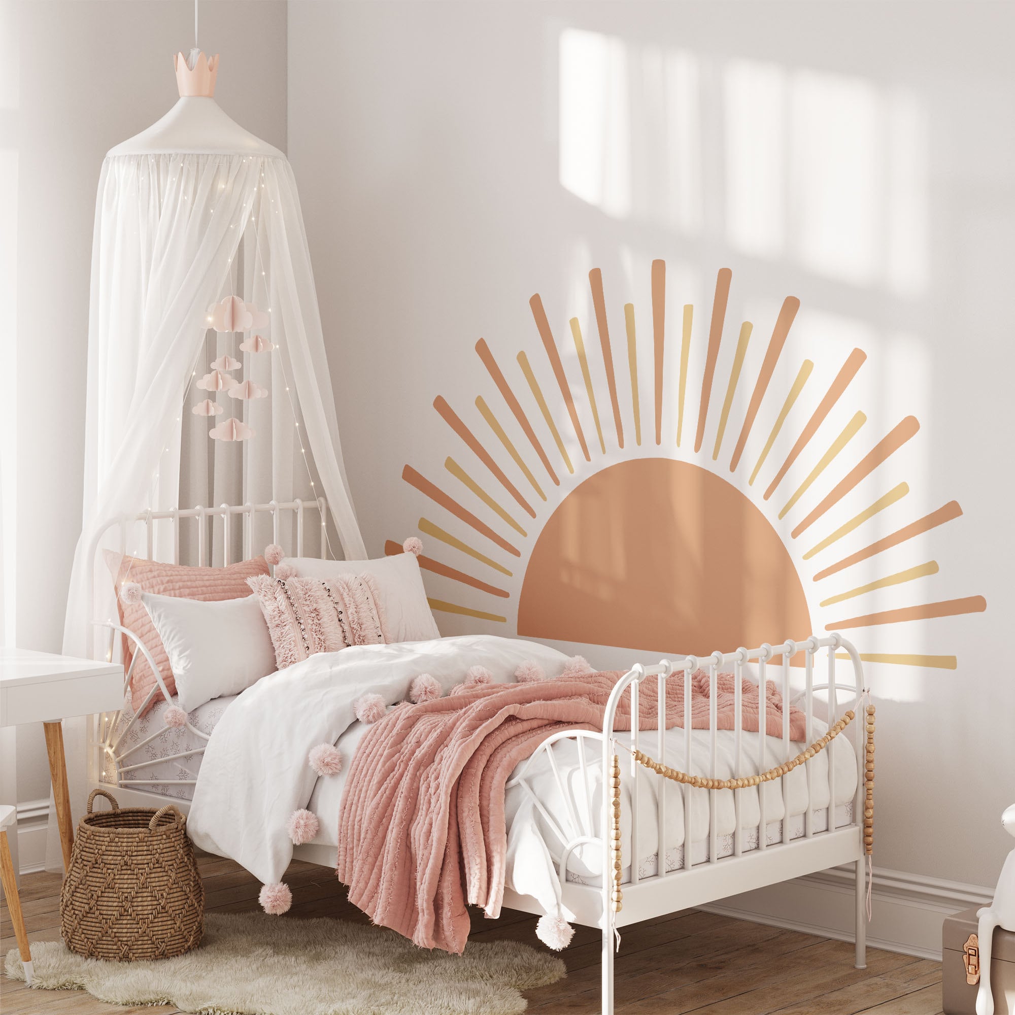 A cozy childs bedroom features a white metal bed with pink and white bedding under a whimsical wall decal. The Boho Sun Sticker by My Store adds charm alongside a canopy with hanging decorations. A wicker basket is on the floor, while natural light enhances the delightful nursery decor.