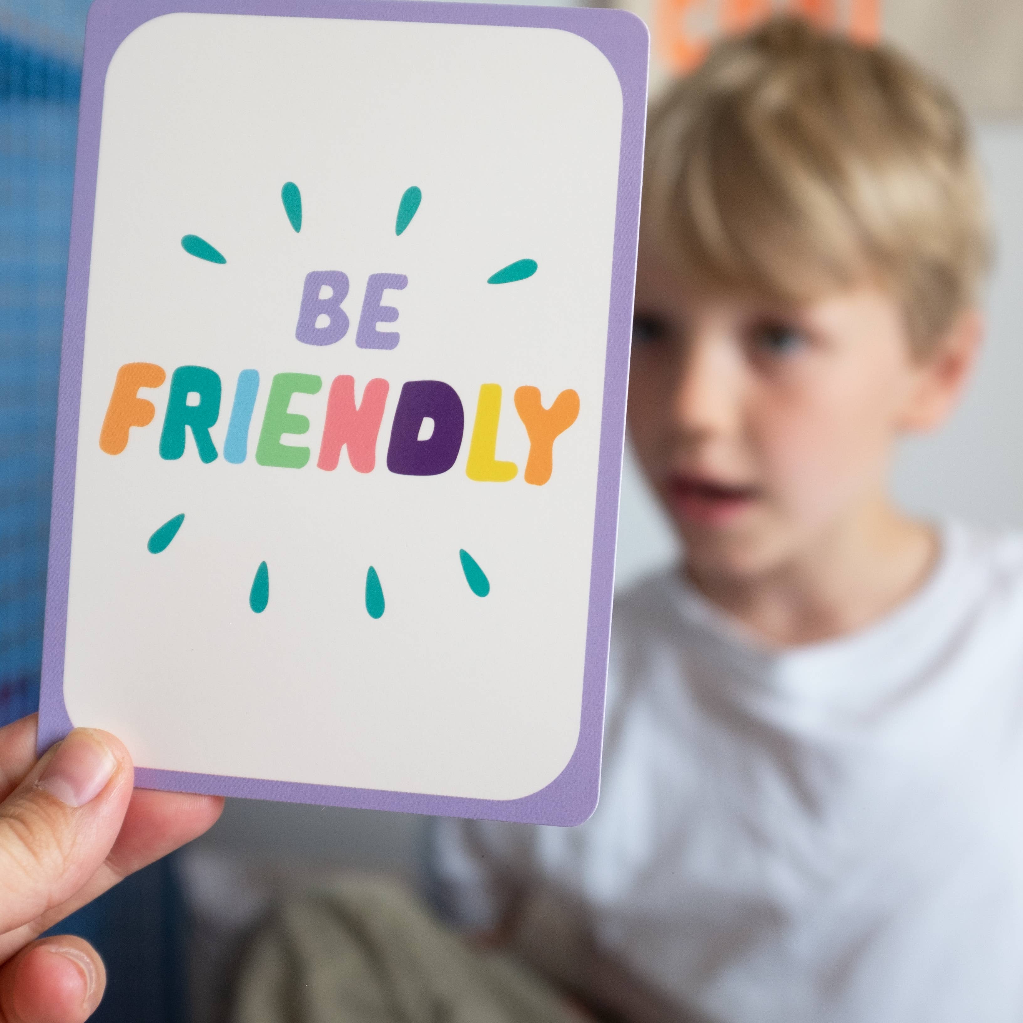 A hand holds a "Be Friendly" card from Mindful Champs' Character Matters Flashcards for Kids, with a child in a white shirt blurred in the background.