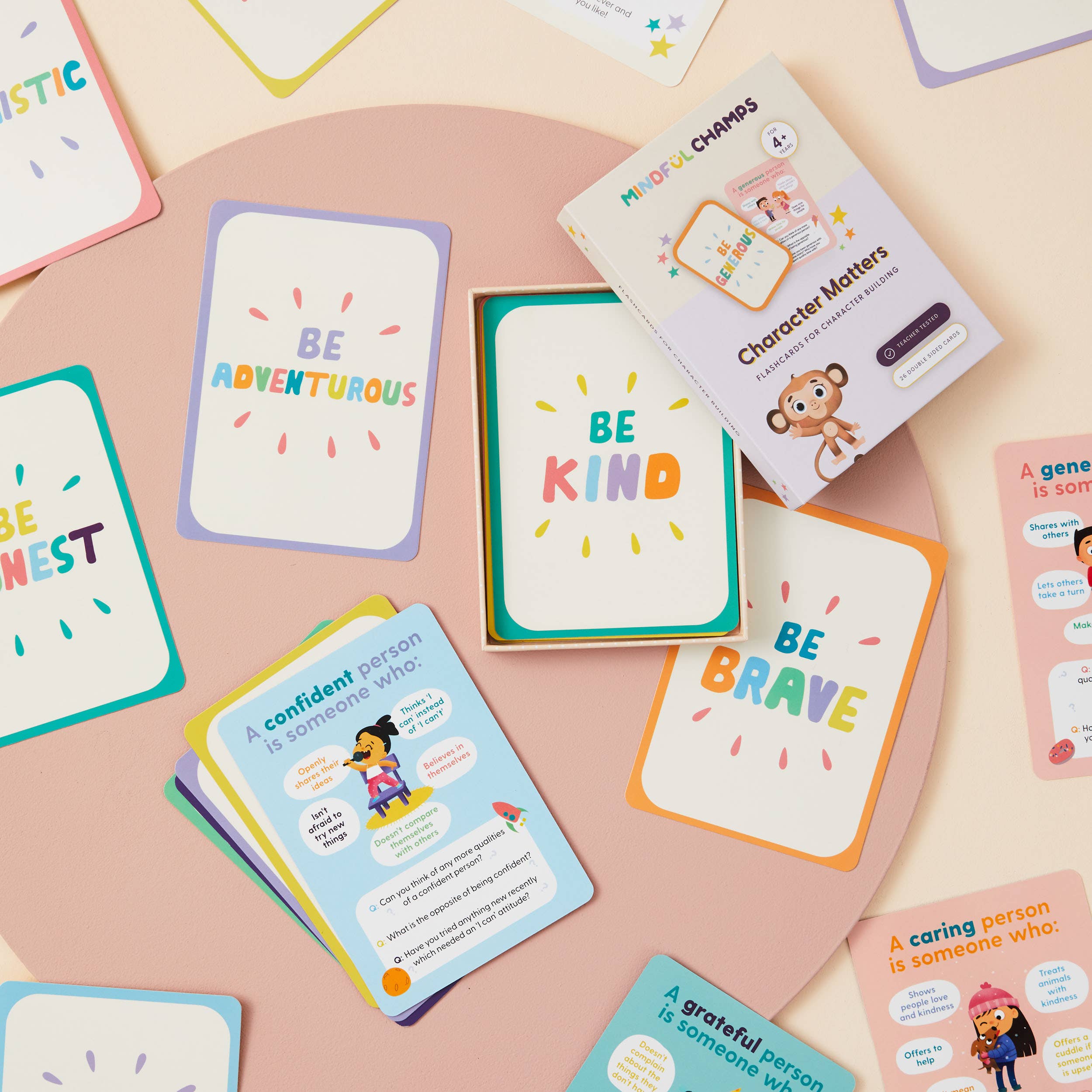 Mindful Champs' Character Matters Flashcards for Kids are displayed on a round pink surface, showing positive messages like Be Adventurous, Be Kind, and Be Brave. The deck's box features a cartoon monkey.