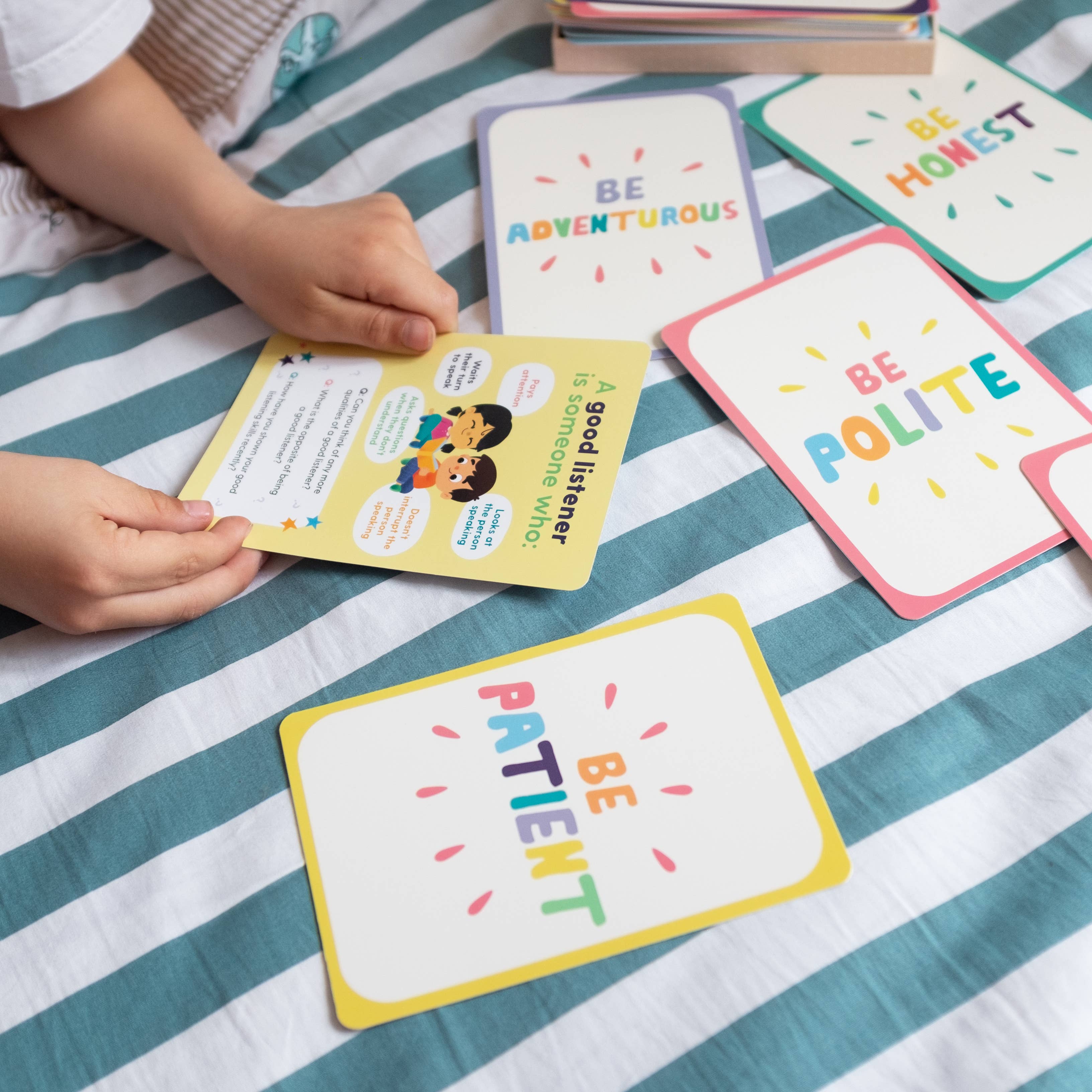 A child sits on a striped bedspread, arranging Mindful Champs' Character Matters Flashcards for Kids with positive messages like Be Adventurous and Be Honest, and holds one about being a good listener to encourage character development through play.