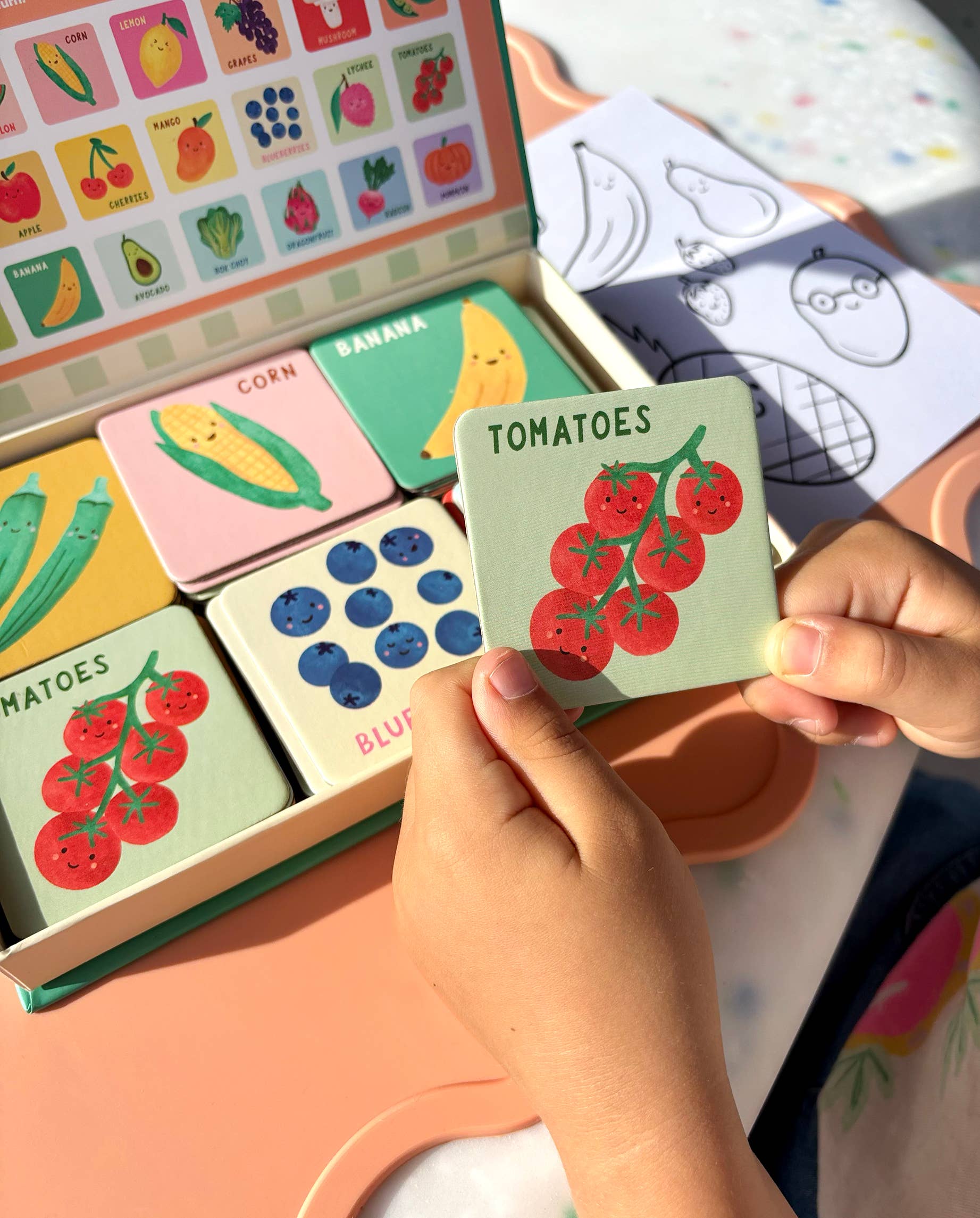 A child holds a card with red tomatoes from the Lauren Sissons Studio Fruit and Veg Memory & Matching Game—Kids Educational Gift. Illustrated cards and the branded box with labeled images are also on the table.