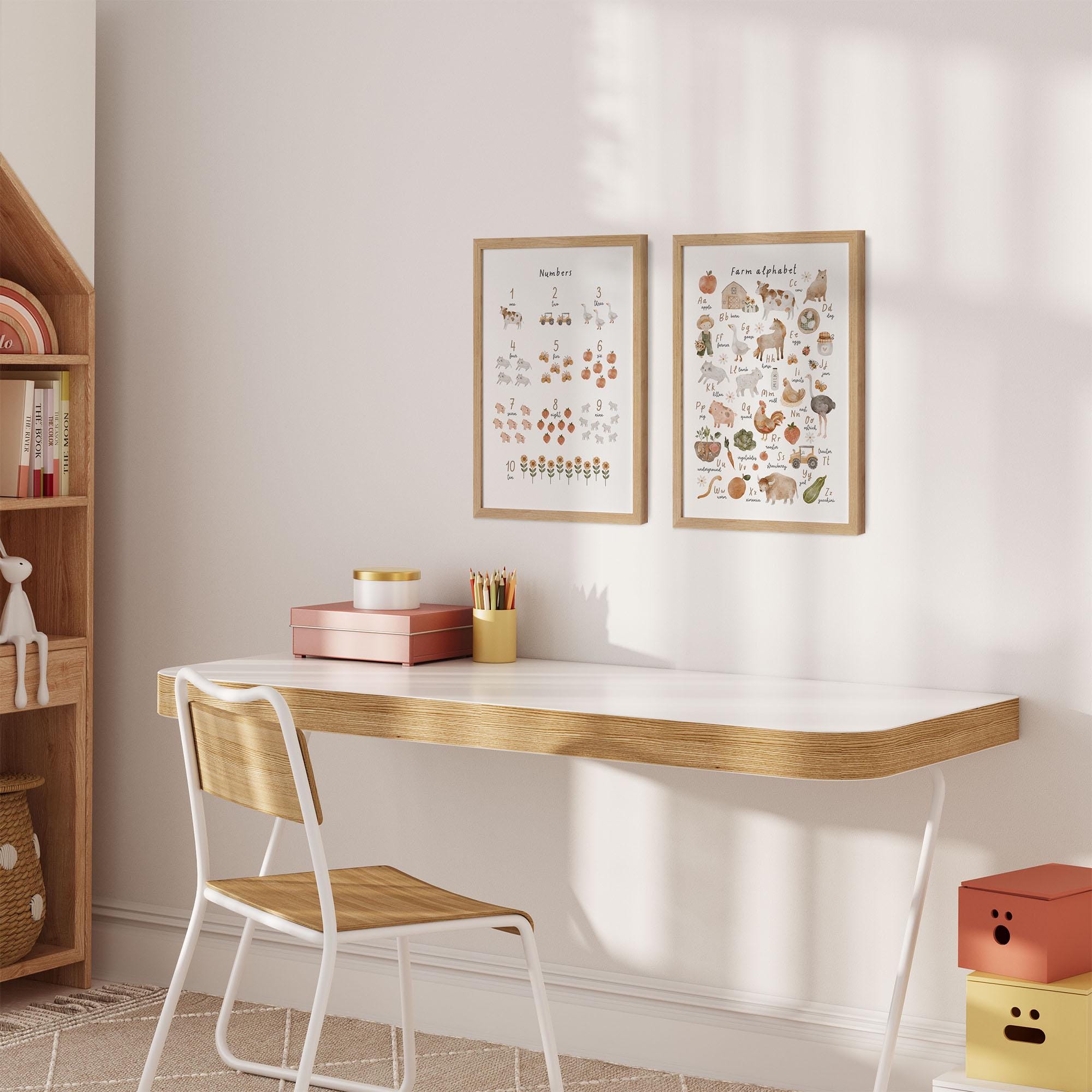A minimalist room showcases a sleek modern wooden desk with white legs, accompanied by a wooden chair and organized stationery. Two framed Farm Animals Alphabet & Numbers nursery prints from My Store adorn the wall. A bookshelf filled with books and a toy occupies the corner.