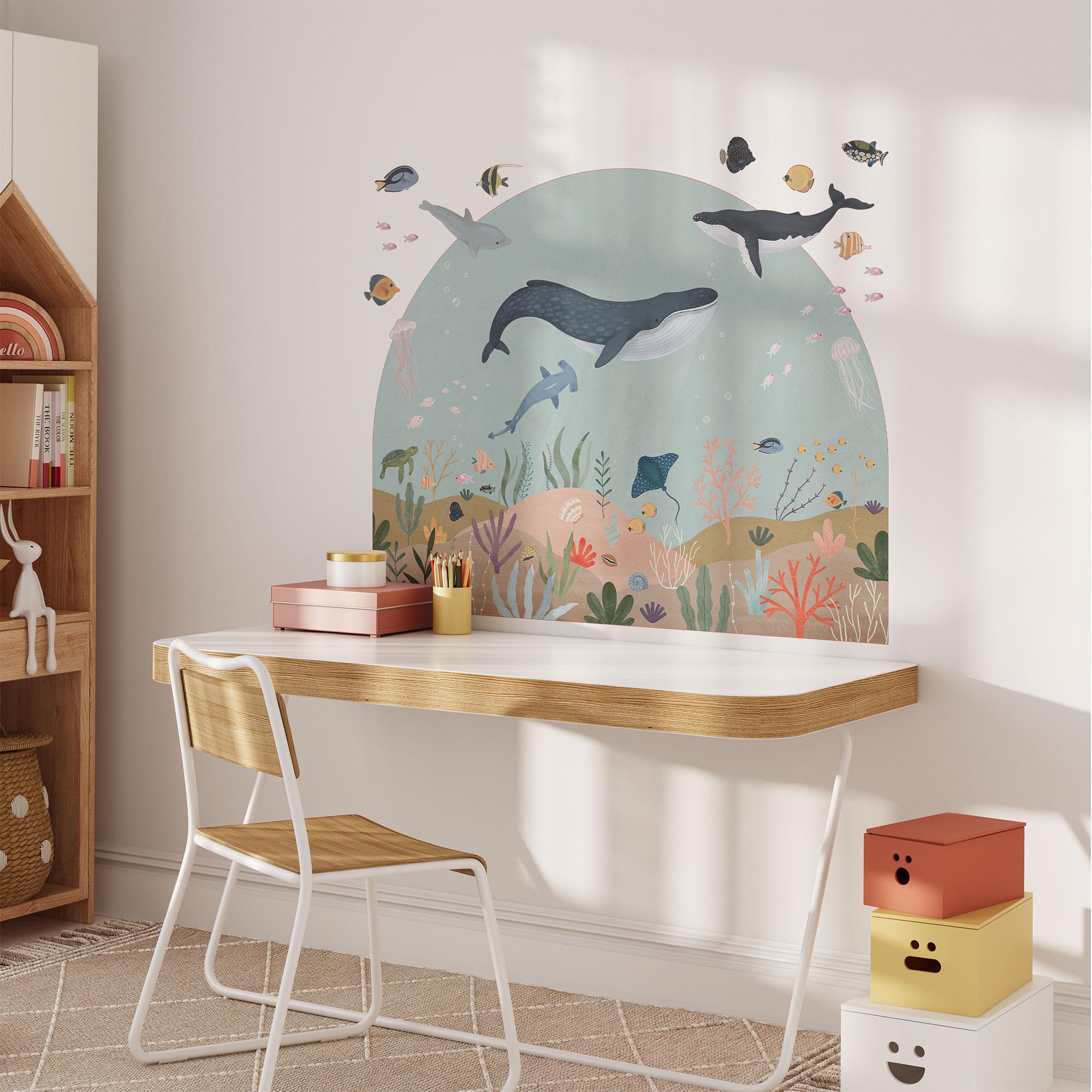 A childs room features a wooden desk and chair, with an ocean-themed mural using My Stores Sea Animals Arch Wall Sticker depicting whales, fish, and coral. Soft sunlight enhances the playful vibe, complemented by colorful stackable drawers on the floor.