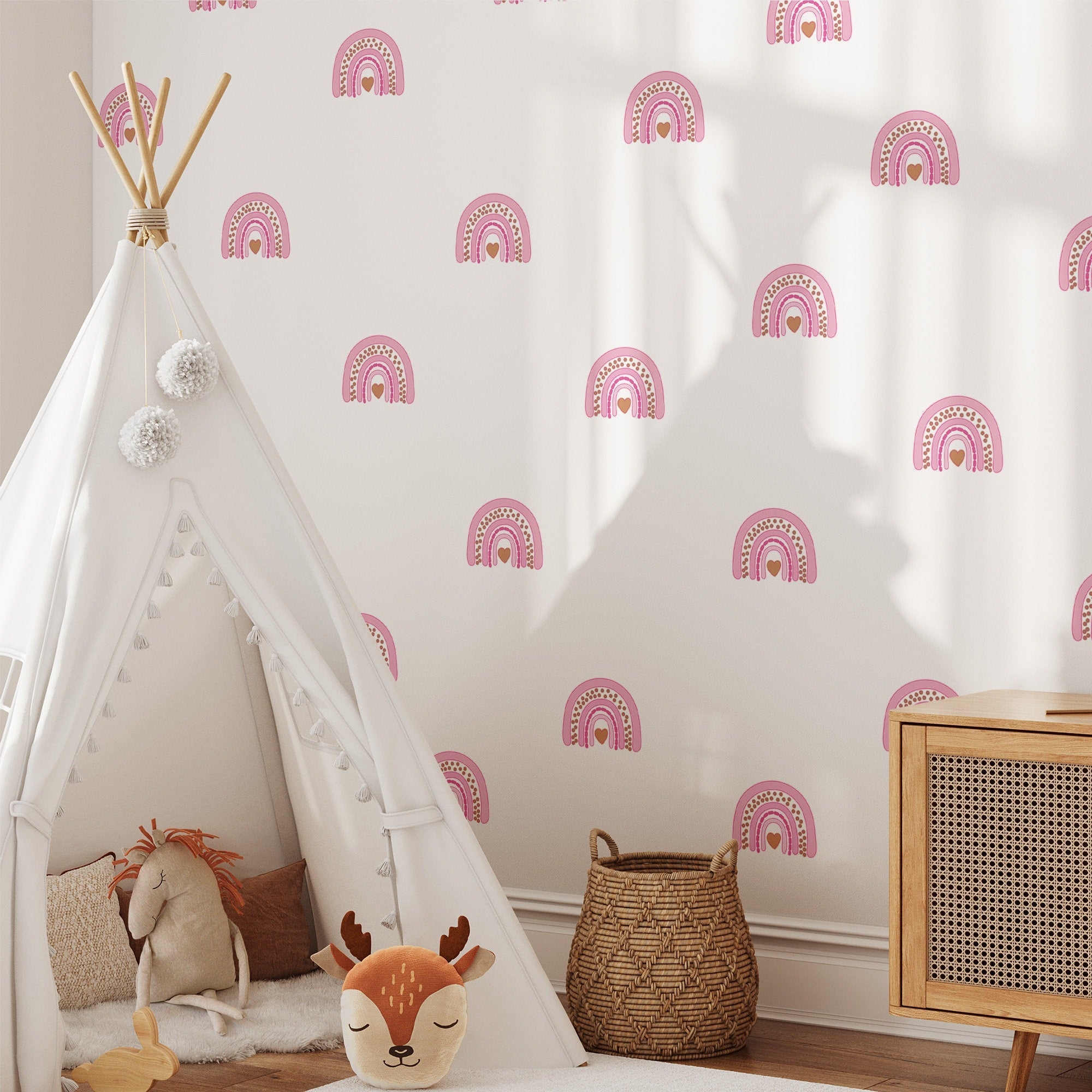 A cozy childrens room includes a teepee with pom-poms, a plush deer toy, and a wicker basket on the floor. The white wall features removable Rainbow Wall Stickers by My Store in pink. A wooden cabinet is partially visible on the right.