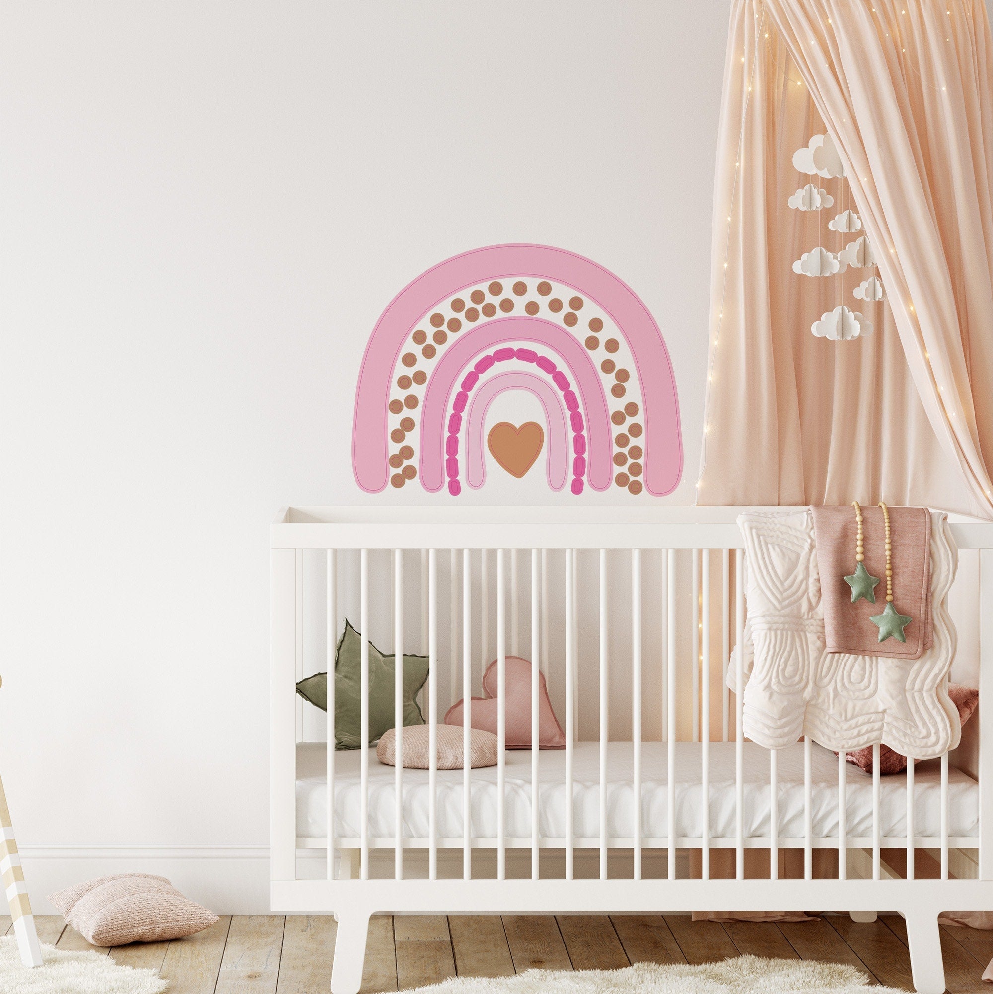 A cozy nursery features a white crib with pink heart and green star pillows, cream bedding, and My Store Rainbow Wall Stickers. Curtains decorated with star string lights and cloud ornaments complete the decor.