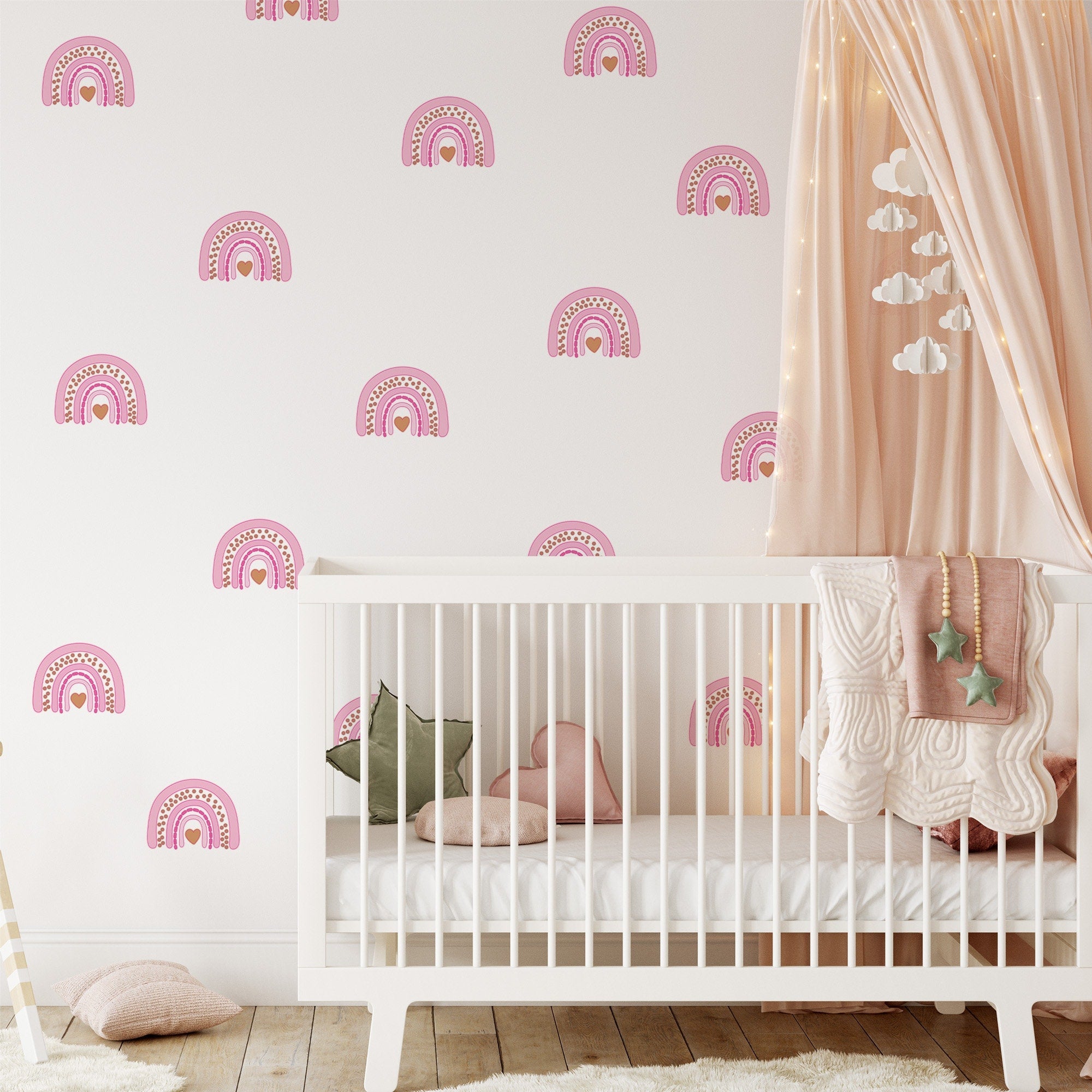 A cozy nursery showcases a white crib with pink and green pillows, draped star garland, and a pink curtain canopy. The wall is adorned with Rainbow Wall Stickers by My Store, completing the rooms warm feel alongside wooden floorboards.