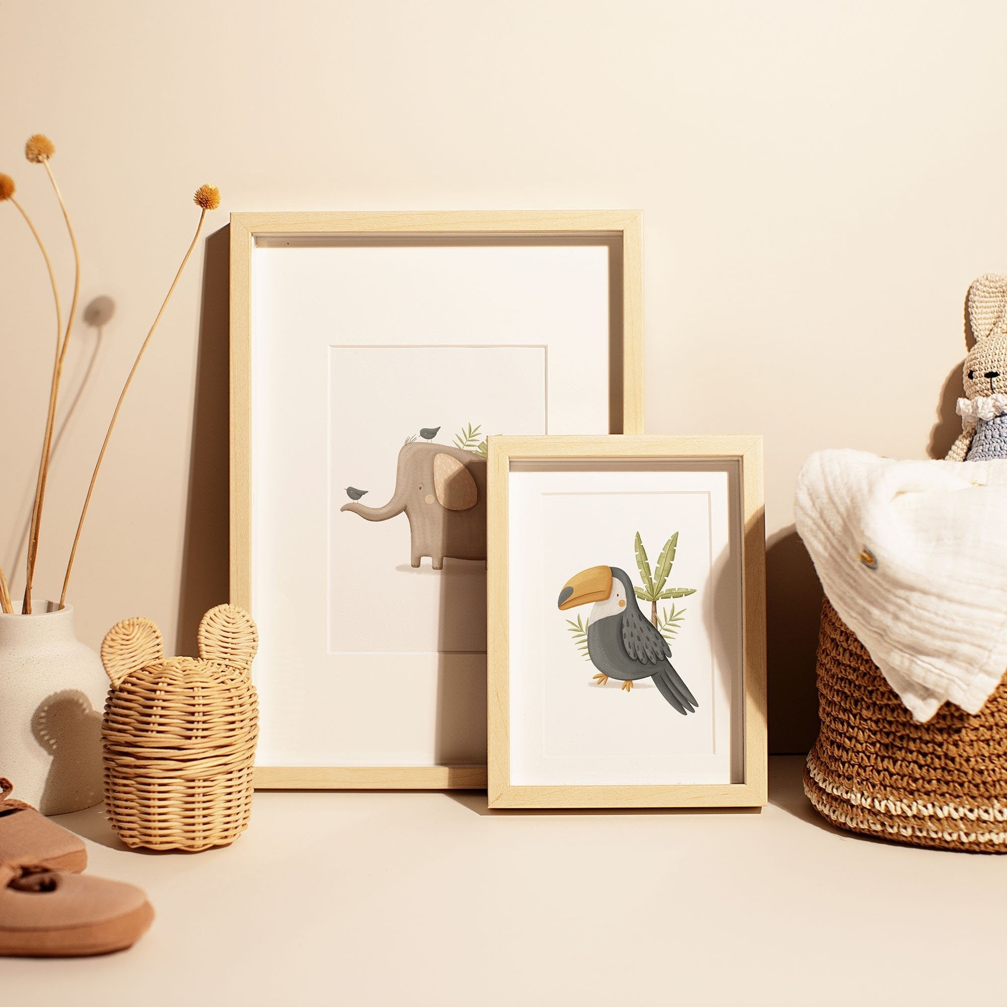 My Stores Safari Animals Nursery Prints, showcasing an elephant and a toucan, adorn a neutral-toned surface. Paired with a woven basket, dried flowers in a vase, and soft baby items, they create a cozy nursery art environment.