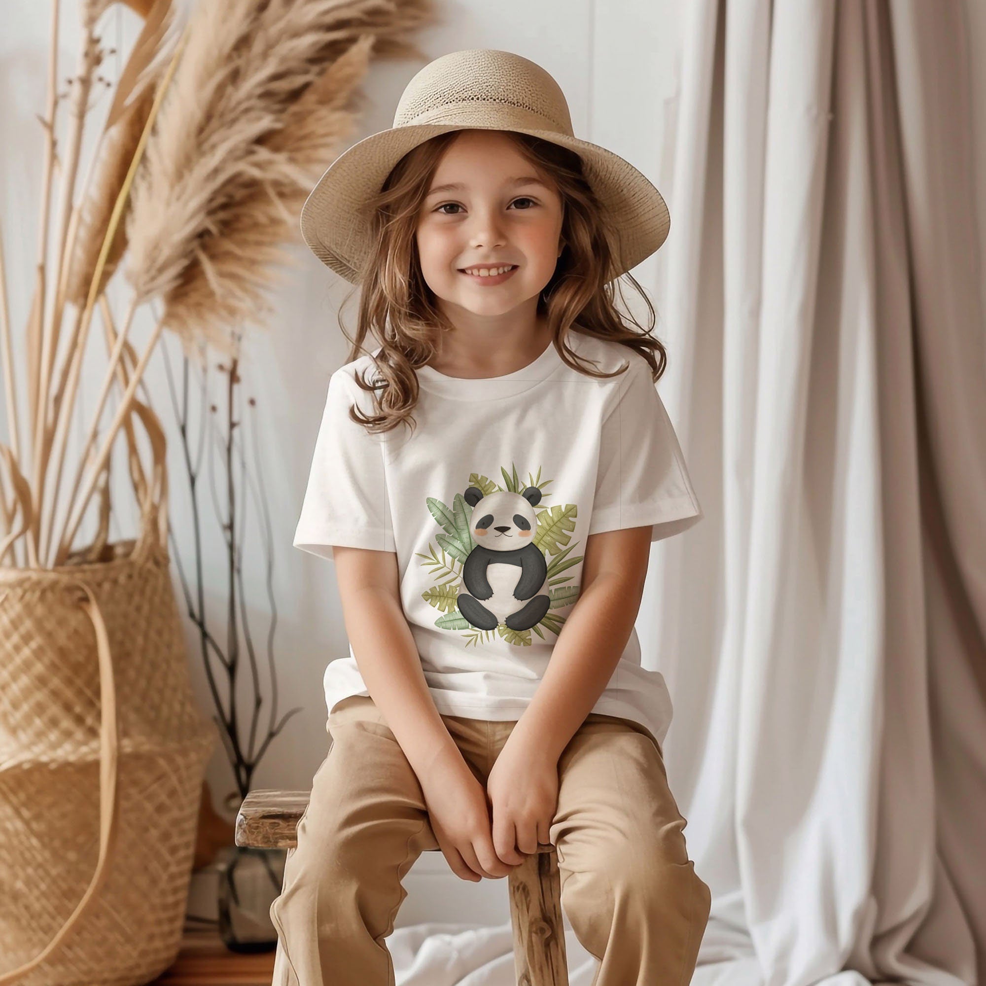 A young girl in a straw hat sits on a wooden stool, smiling warmly in beige pants and surrounded by indoor plants and dried pampas grass. She wears a comfy, stylish Safari Animals Toddler T Shirt from My Store made of 100% ringspun combed cotton, which is machine washable.