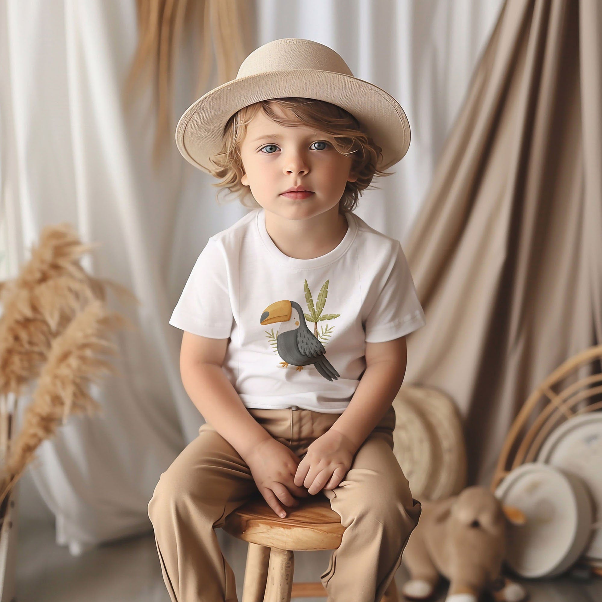 A young child with curly hair sits on a wooden stool, wearing a white Safari Animals Toddler T-Shirt from My Store, made of 100% ringspun combed cotton. The beige pants and straw hat match the neutral-tone drapery and pampas grass backdrop. The outfit is conveniently machine washable.