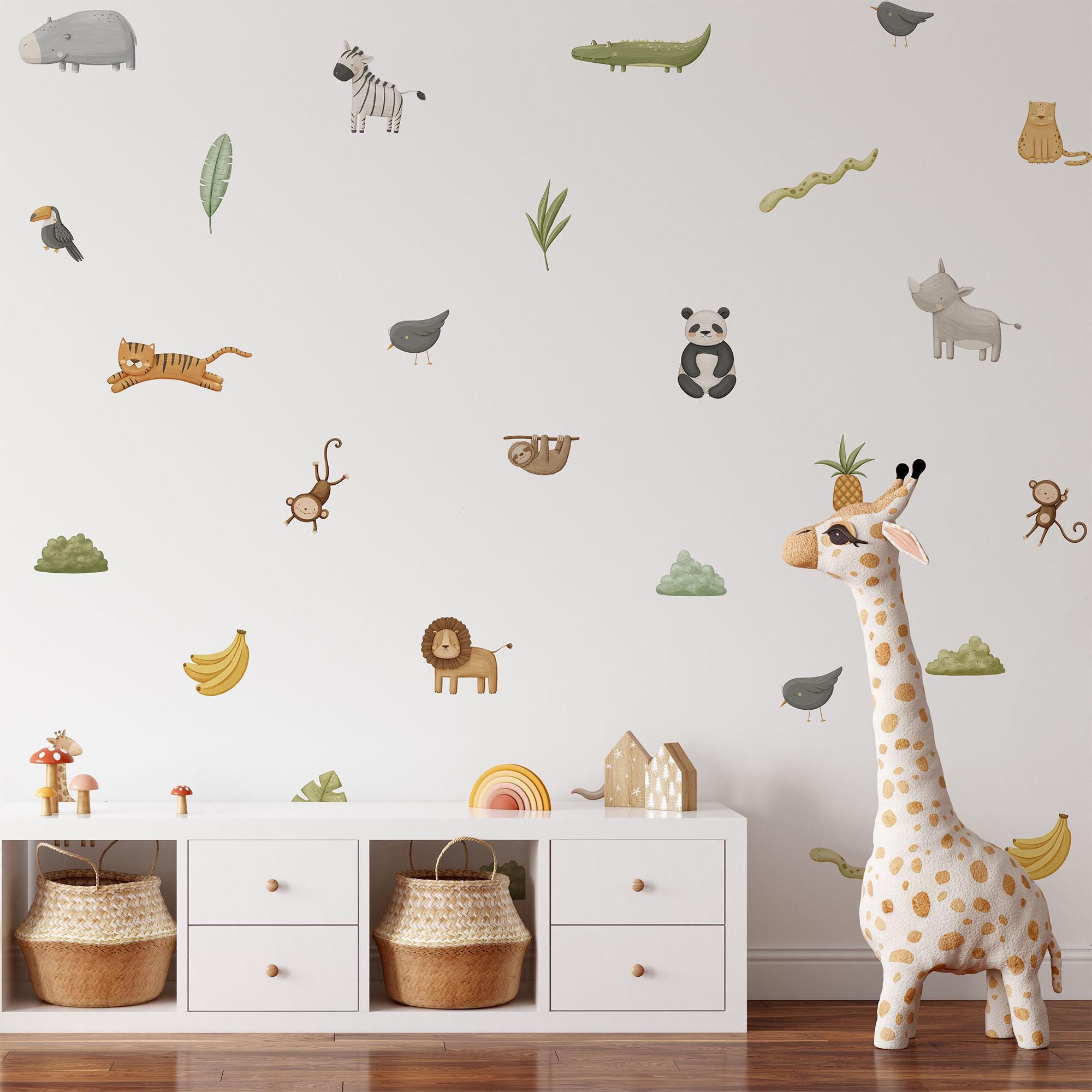 The nursery room displays My Stores Safari & Jungle Animal Stickers, including zebra, lion, panda, and giraffe decals on a white wall. A plush giraffe toy is on the floor next to a white cabinet under the charming stickers.