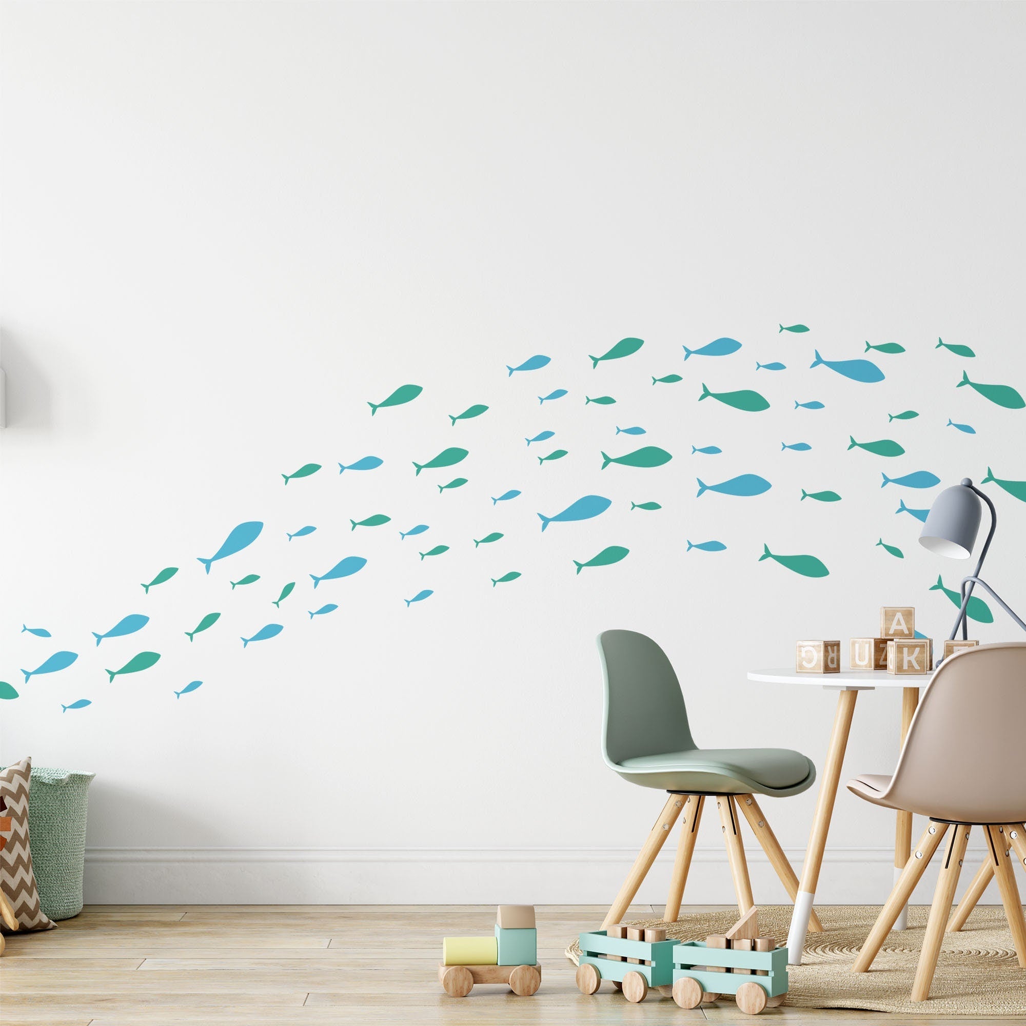 A modern childrens room with wooden floors features white walls adorned with Shoaling Fish Stickers by My Store in teal and blue. The nursery has a small round table, two chairs, a wooden toy car, and a lamp. Wood blocks spell JULK.