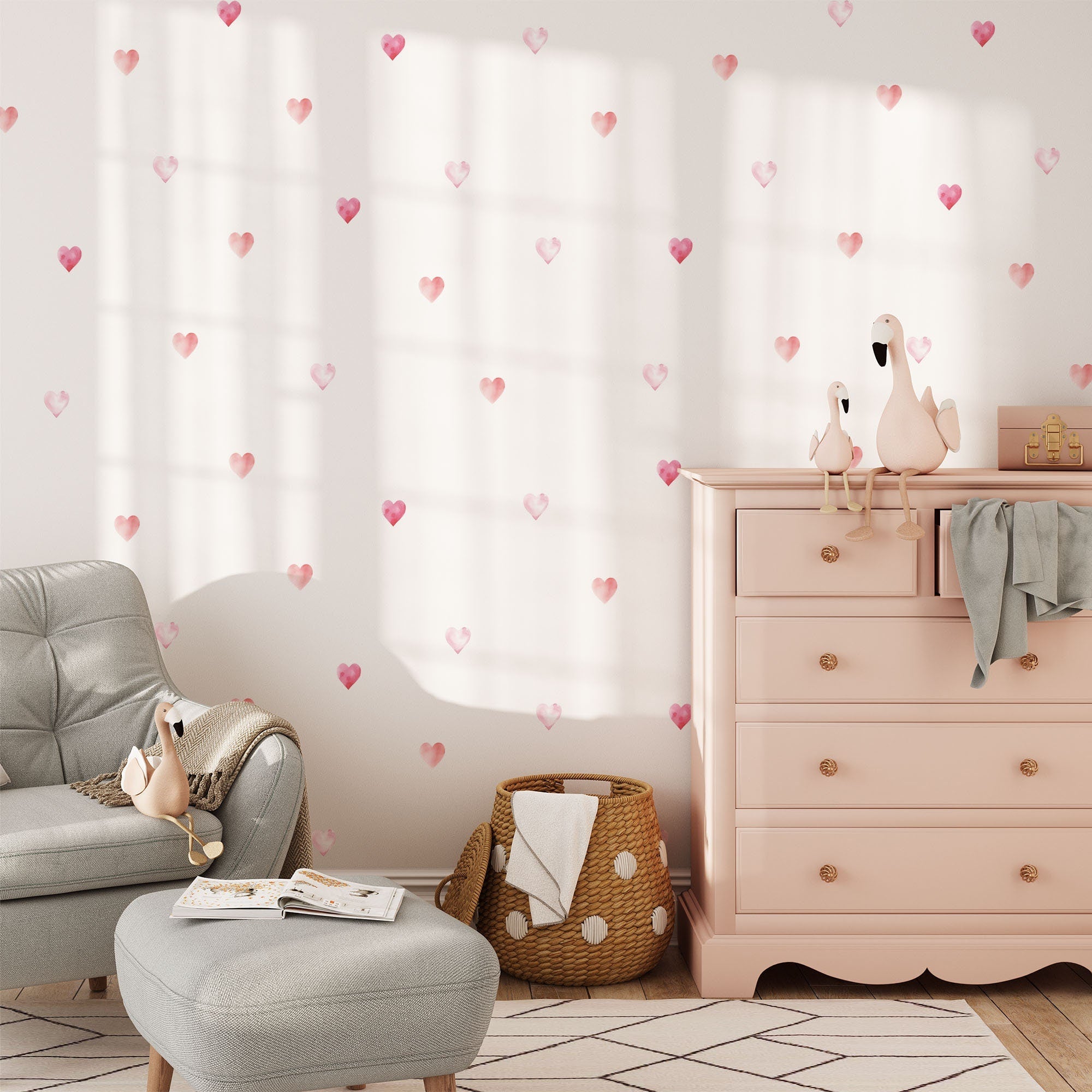 A cozy room includes a light pink dresser with two flamingo figurines. Walls display My Stores Watercolour Heart Stickers. A gray armchair and woven basket complete the space, all gently illuminated by natural light.