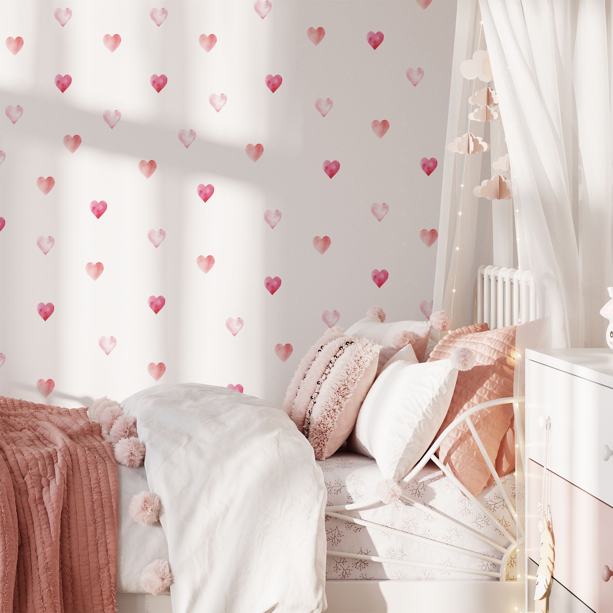 A cozy bedroom showcases Watercolour Heart Stickers by My Store, featuring heart-patterned wallpaper, a white metal bed with pink and white pillows, a chunky pink throw, and fairy lights. Cloud decorations hang above, completing the warm and whimsical look with easily removable decals.