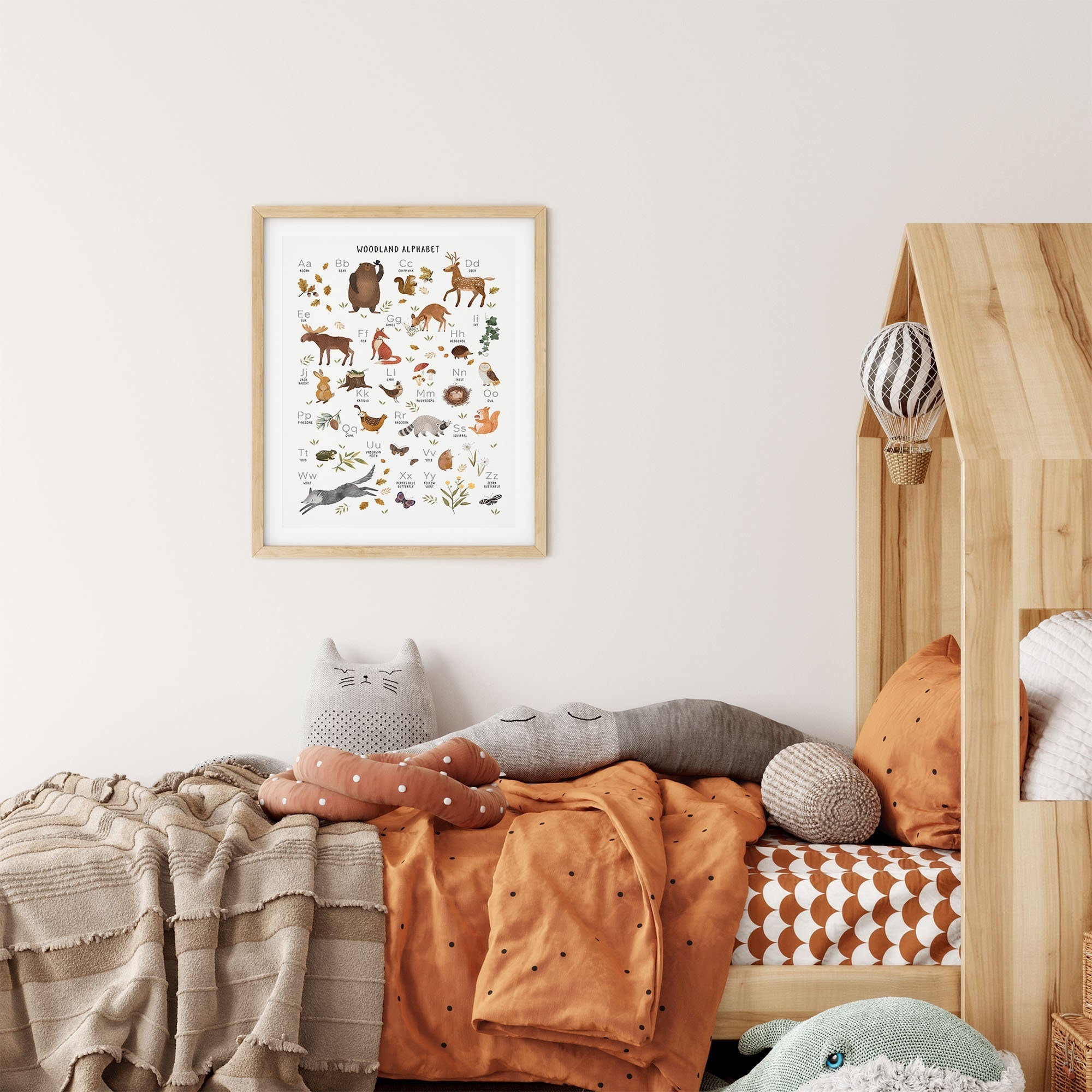 A cozy childrens room includes a wooden bed with orange and beige bedding, adorned with plush cat and crocodile toys. Above hangs the Woodland Alphabet Nursery Print by My Store, providing charming nursery art that complements the soft, neutral color palette.