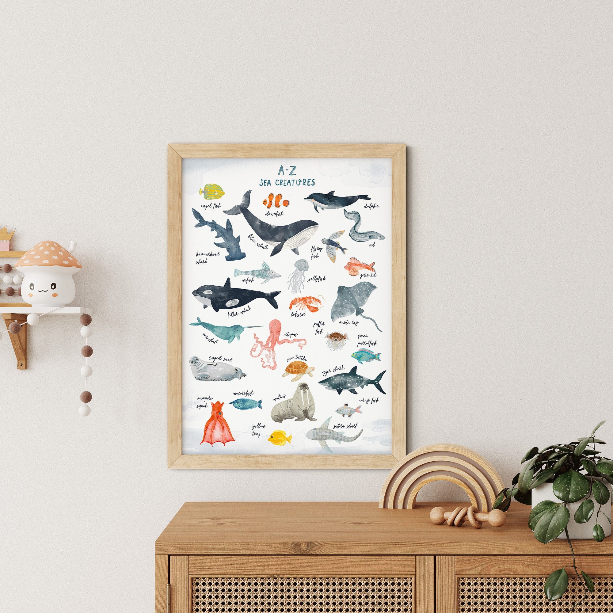 The My Store A-Z Sea Creatures Nursery Print, framed and educational, features illustrated sea animals with names. Its perfect nursery art hanging on a light wall above a wooden cabinet, with a nearby shelf holding a small pot and decorative item.