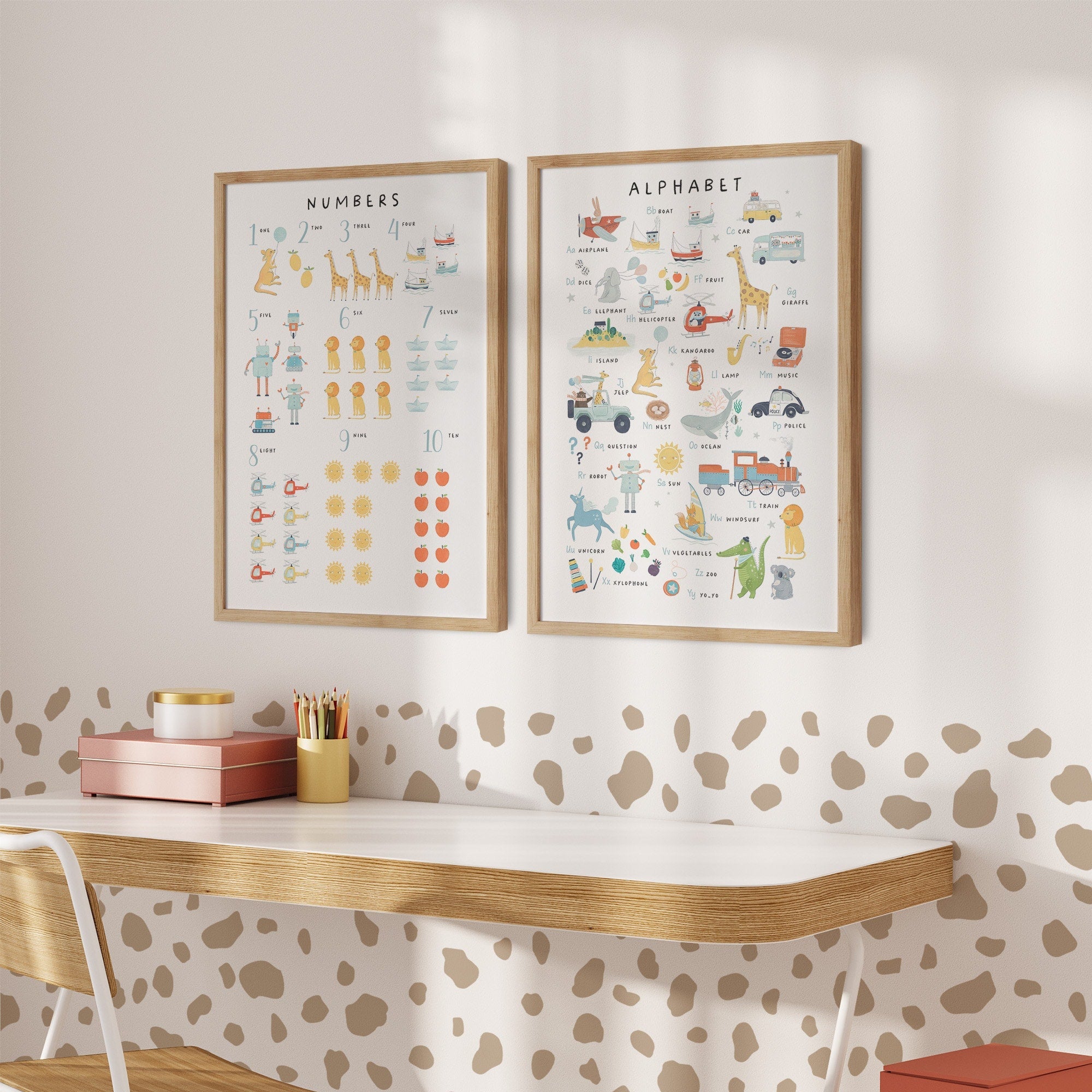 A childrens room features a wooden desk, white chair, and My Stores Numbers & Alphabet Nursery Print. One poster displays numbers with illustrations, the other showcases the alphabet with images. Warm lighting and a spot-patterned wall complete the playful decor.