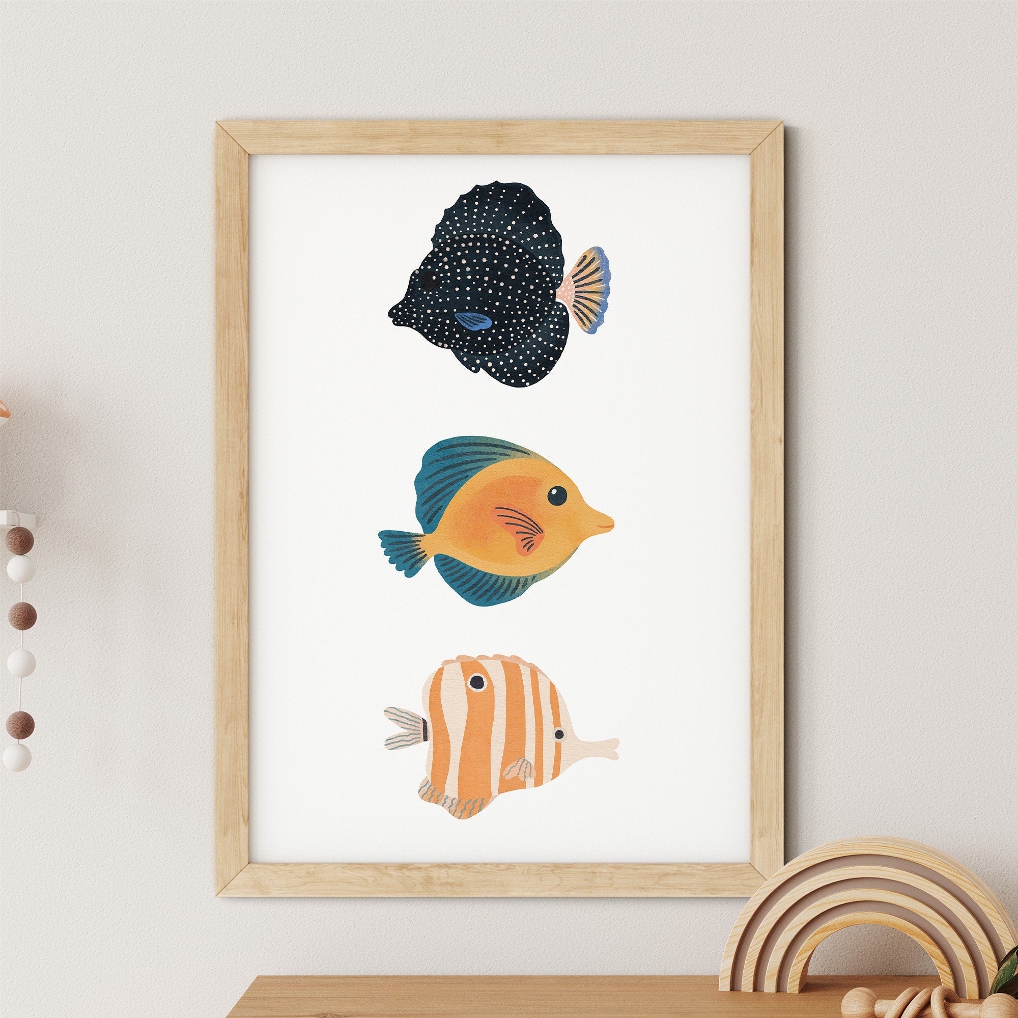 The Reef Fish Nursery Print by My Store showcases three charming reef fish illustrations— a dotted dark blue fish, an orange and blue fish, and a striped orange & white fish—on a white background in a wooden frame, making it ideal nursery art with subtle decor on the side.