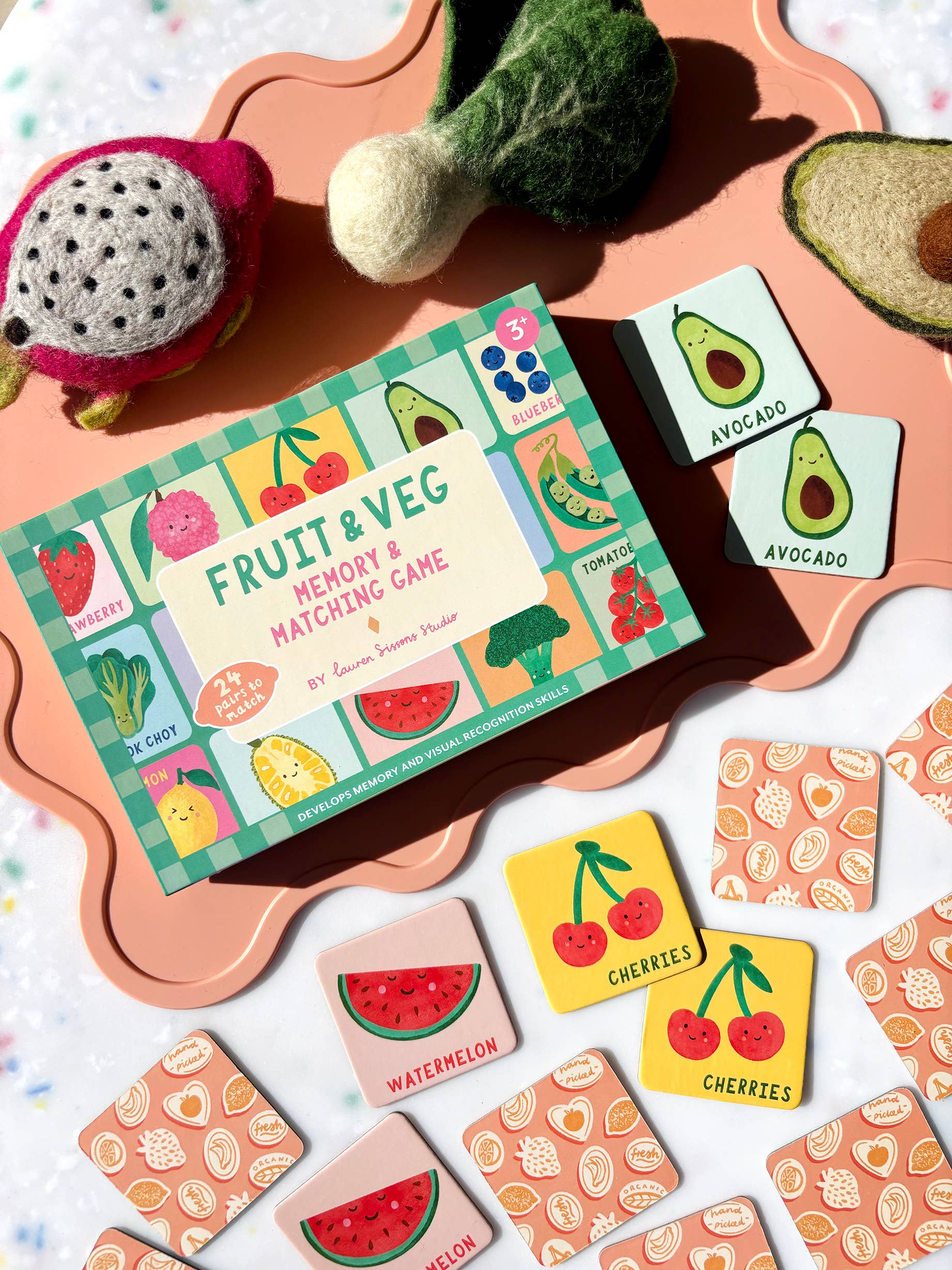 A fruit and vegetable-themed memory and matching game with illustrated cards showing items like avocado, watermelon, and cherries, displayed on a peach tray with felt fruit toys nearby.