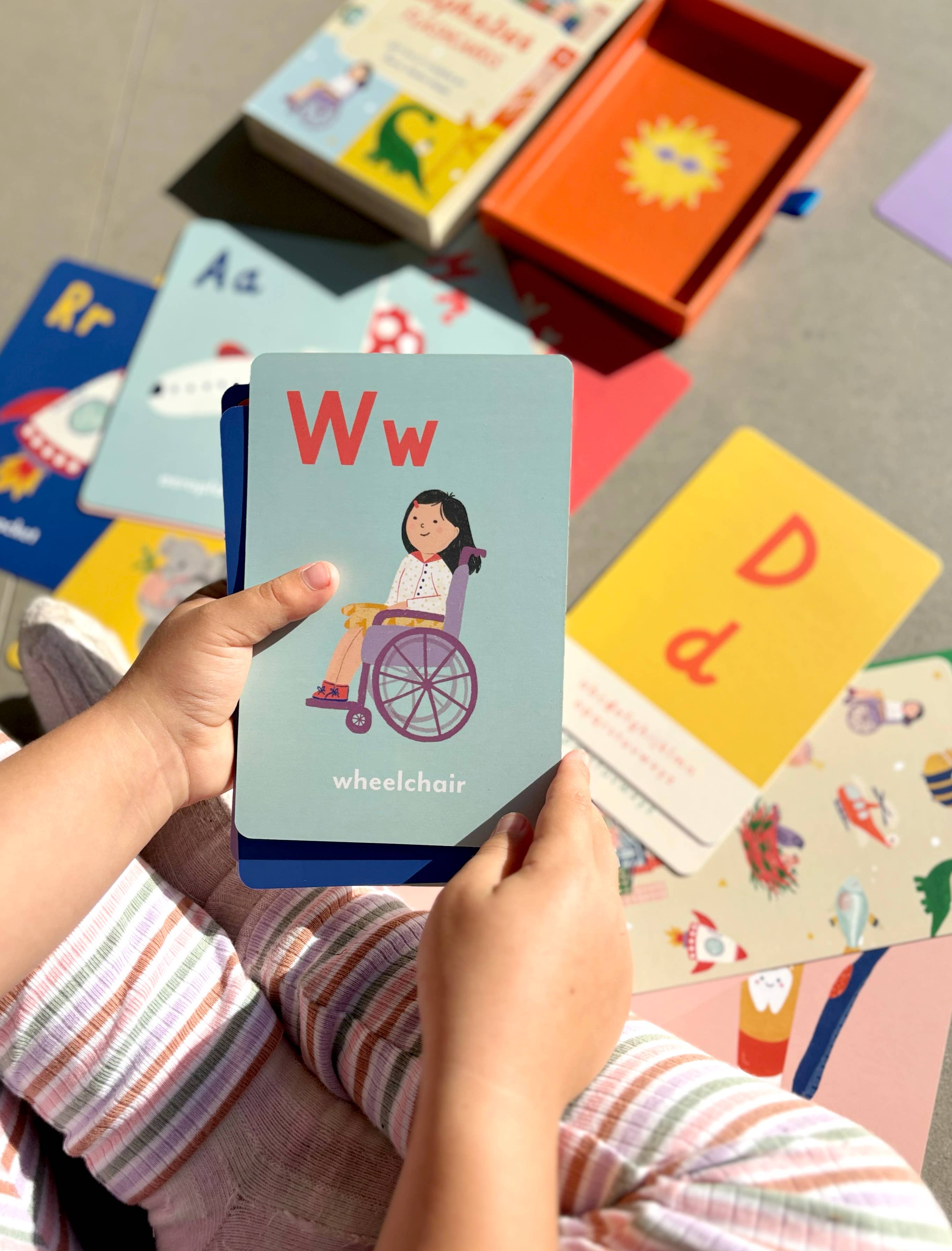 A child holds a flashcard showing the letter W with an illustration of a girl in a wheelchair. Colorful alphabet flashcards and books are spread out on the floor in the background.