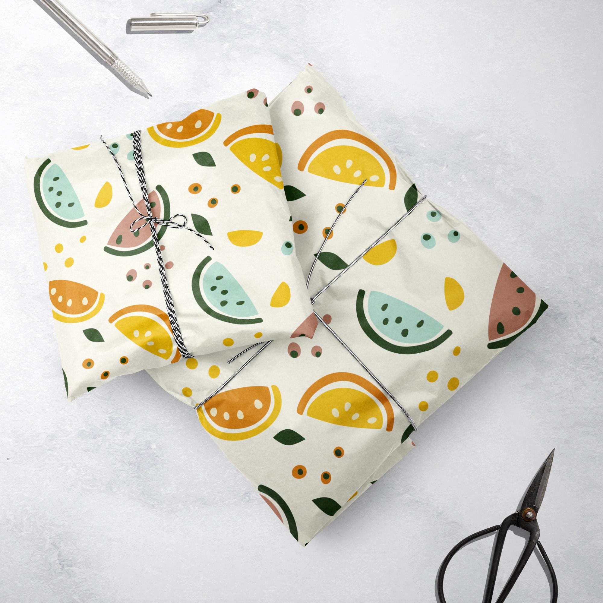 Two gifts wrapped in white paper with colorful fruit patterns, including oranges and watermelons, tied with black-and-white string. Scissors and a pen are nearby on a light-colored surface.