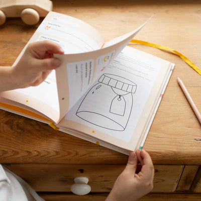 A child flips through the My Mindful Me Journal by Mindful Champs on a wooden desk, discovering a page with a jar illustration. A pencil and wooden toy nearby encourage kids’ mental health and daily self-reflection.