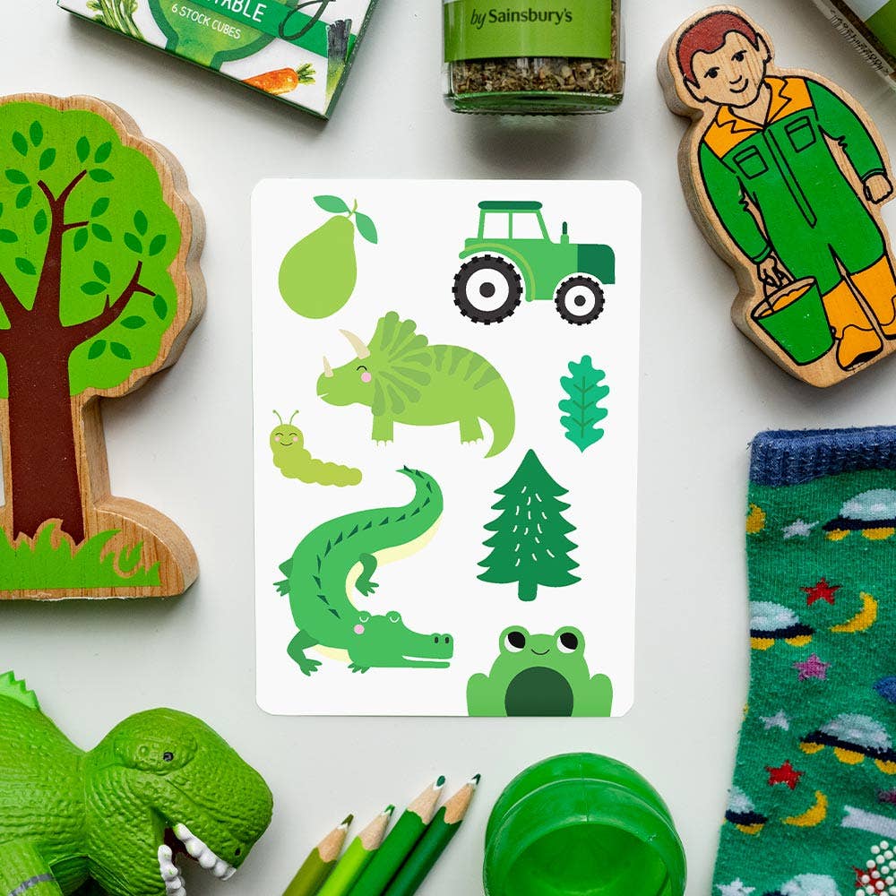 A flat lay of mostly green children's toys and objects surrounds a Priya & Peanut Colour Flash Card featuring green items like a pear, tractor, triceratops, leaf, caterpillar, crocodile, tree, and frog—ideal for early learning.