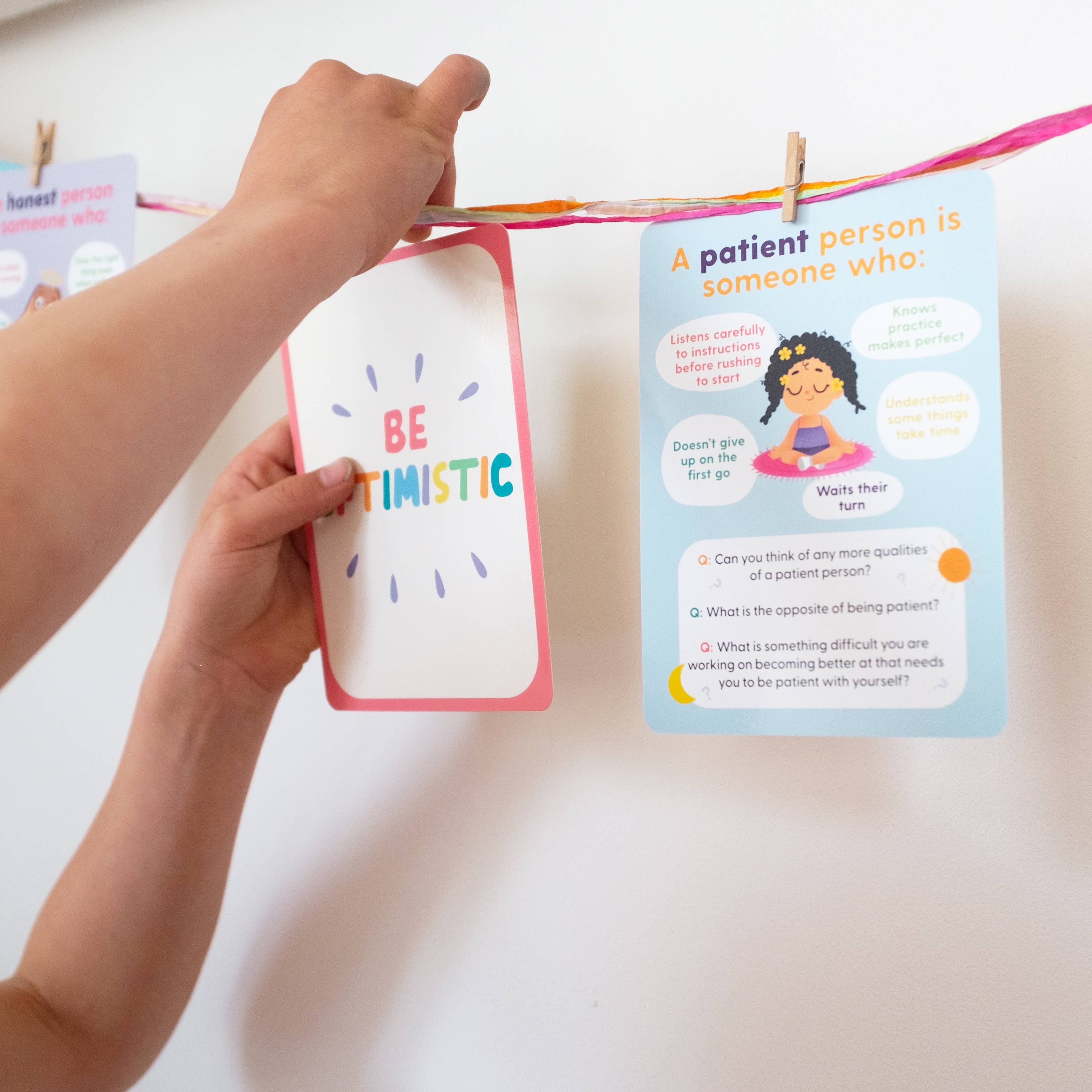 A person hangs a "BE OPTIMISTIC" card from the Mindful Champs Character Matters Flashcards for Kids beside another card about developing patience, both clipped to a line with more motivational flashcards in the background.
