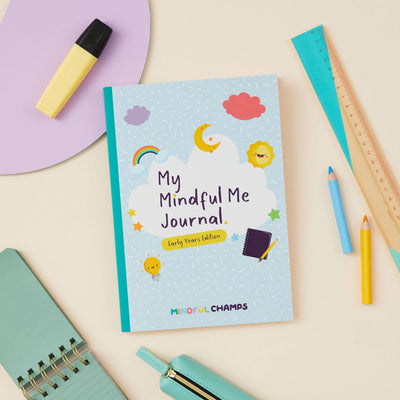 The My Mindful Me Journal Early Years Edition by Mindful Champs rests on a pale yellow desk, surrounded by a ruler, pencils, highlighter, notebook, and a zipped mint pencil case.