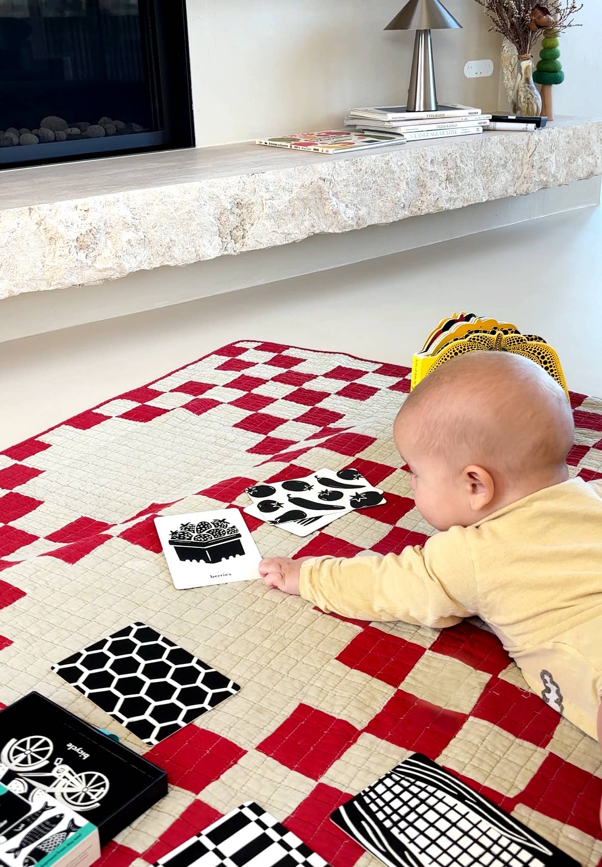 A baby lies on a red and white checkered quilt, reaching for Lauren Sissons Studio's High Contrast Flashcards for Baby—sensory newborn gift cards designed to support early visual development. A stone shelf and various objects appear in the background.