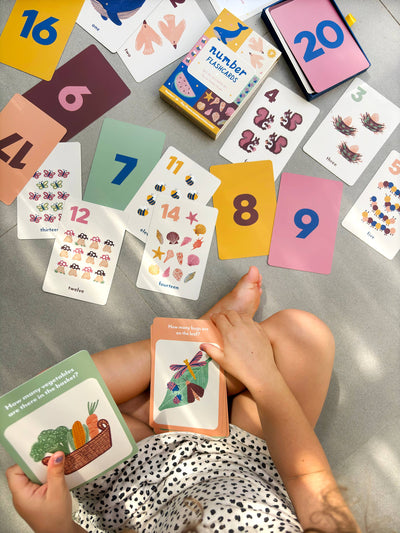 A child sits on the floor holding Lauren Sissons Studio’s Illustrated Number Flashcards - Kids Toys Learning Education, surrounded by colorful cards featuring numbers, animals, and objects, plus a blue and yellow flashcard box.