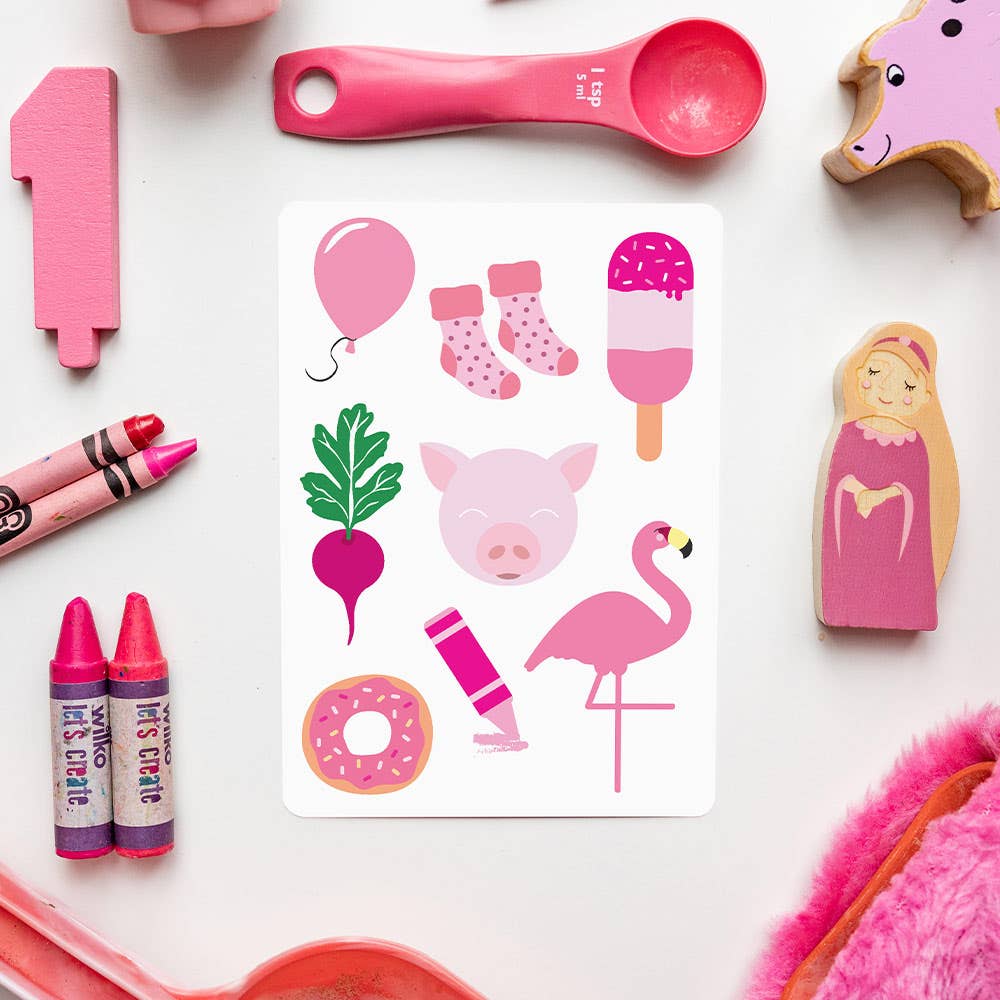 A Priya & Peanut Colour Flash Card featuring pink illustrations—pig, flamingo, balloon, ice cream, donut, crayon, beet, socks, and heart—is surrounded by pink toys and objects on a white surface.