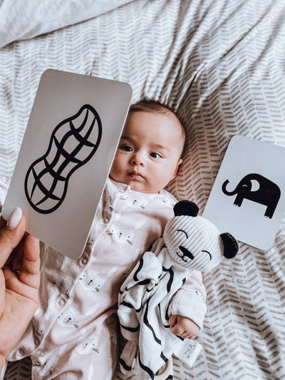 A baby on a patterned blanket holds a soft panda toy. An adult shows Priya & Peanut Newborn Sensory Cards (0+ Months Old) with a peanut illustration, while an elephant card from the award-winning set rests nearby.